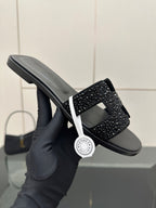 Black slide sandal held in gloved hand, rhinestone strap across top
