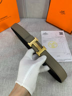 Olive leather belt with gold H buckle, held in white-gloved hand