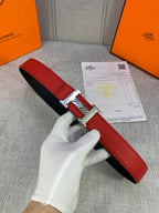 Red leather belt with chrome H buckle, shown flat on a gray fabric surface.