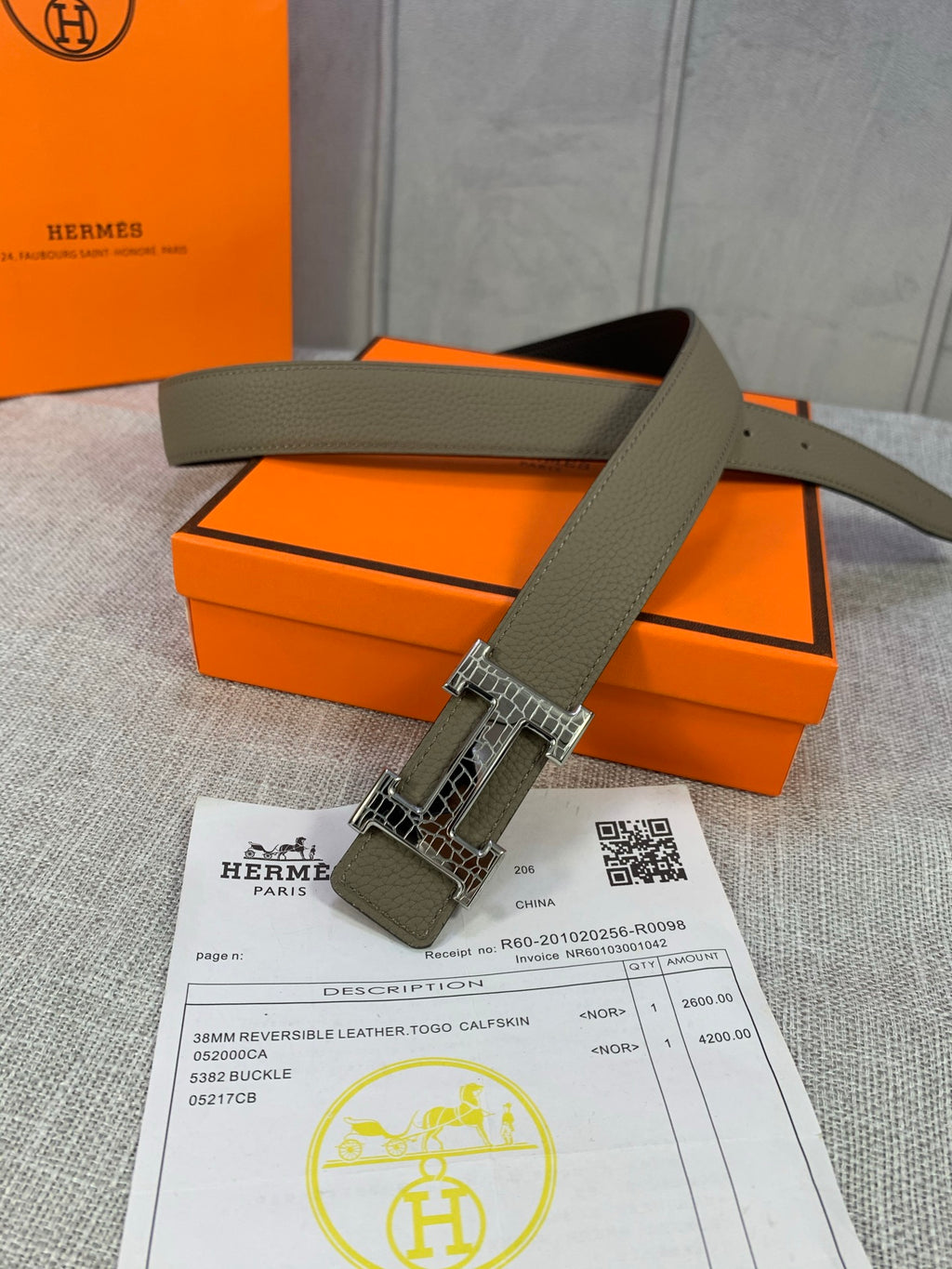 Olive leather belt with silver buckle and rectangular keeper tab, laid flat on box.
