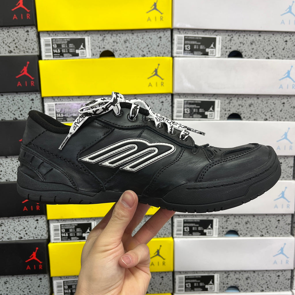 Black low-top sneaker front view with silver logo patch and laces