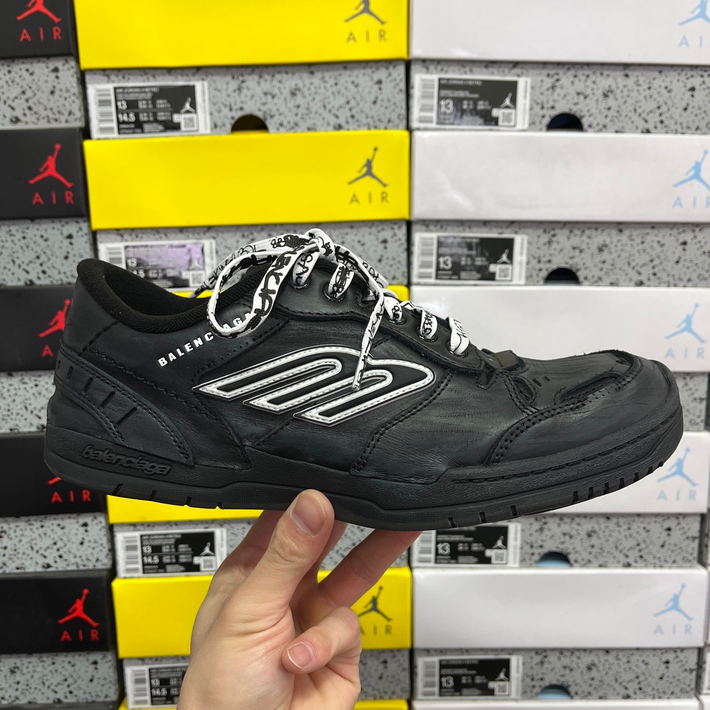 Black low-top sneaker with silver logo patch and lace-up upper