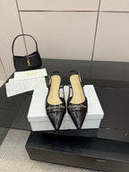 Black pointed-toe slingback heels with gold logo detail, shown on display boxes