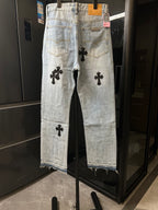 Light wash denim pants with black cross patches on front legs, button fly