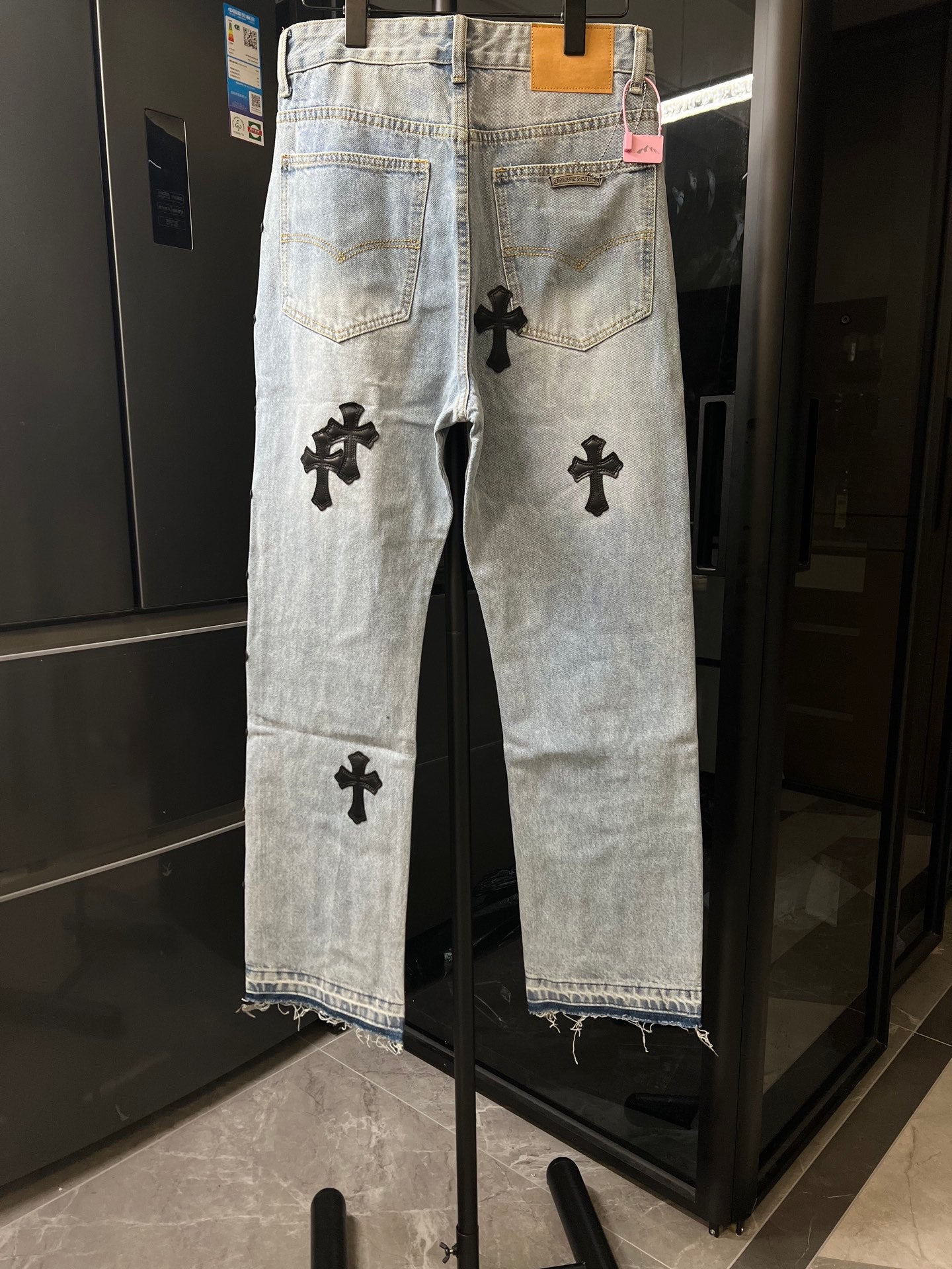 Light wash denim pants with black cross patches on front legs, button fly