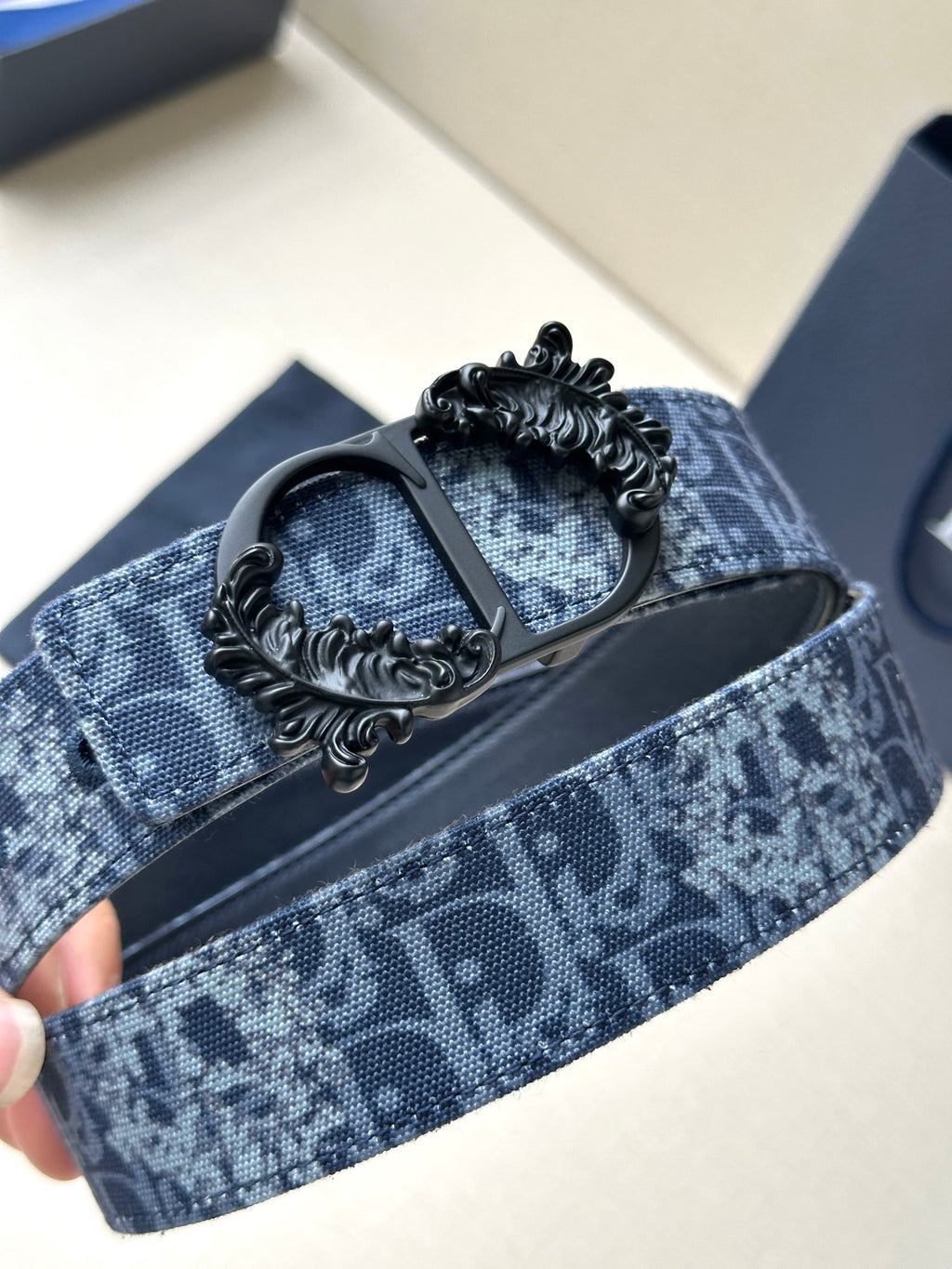Blue monogram belt with black strap backing and detailed ornate buckle centerpiece.