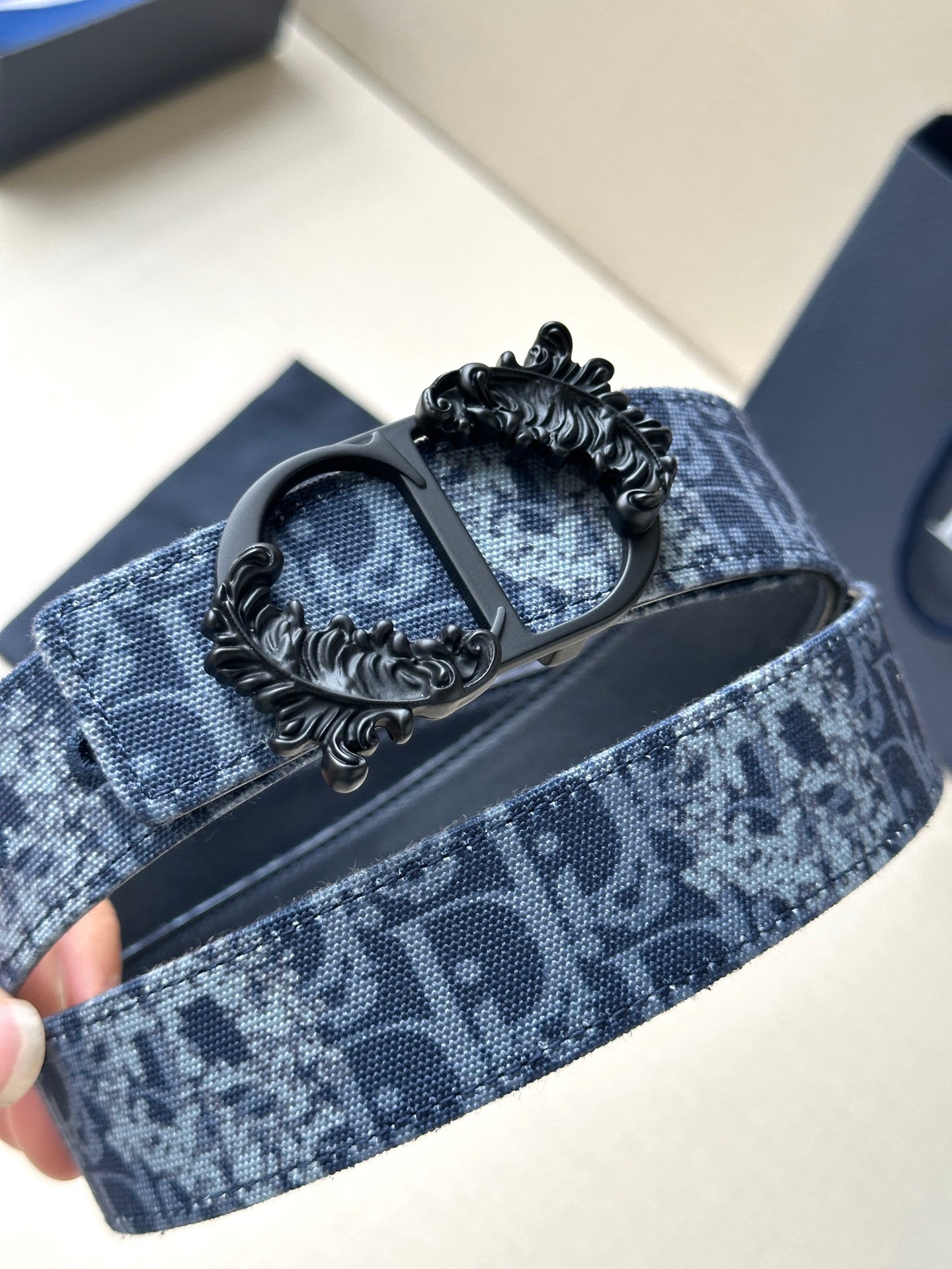 Blue monogram belt with black strap backing and detailed ornate buckle centerpiece.