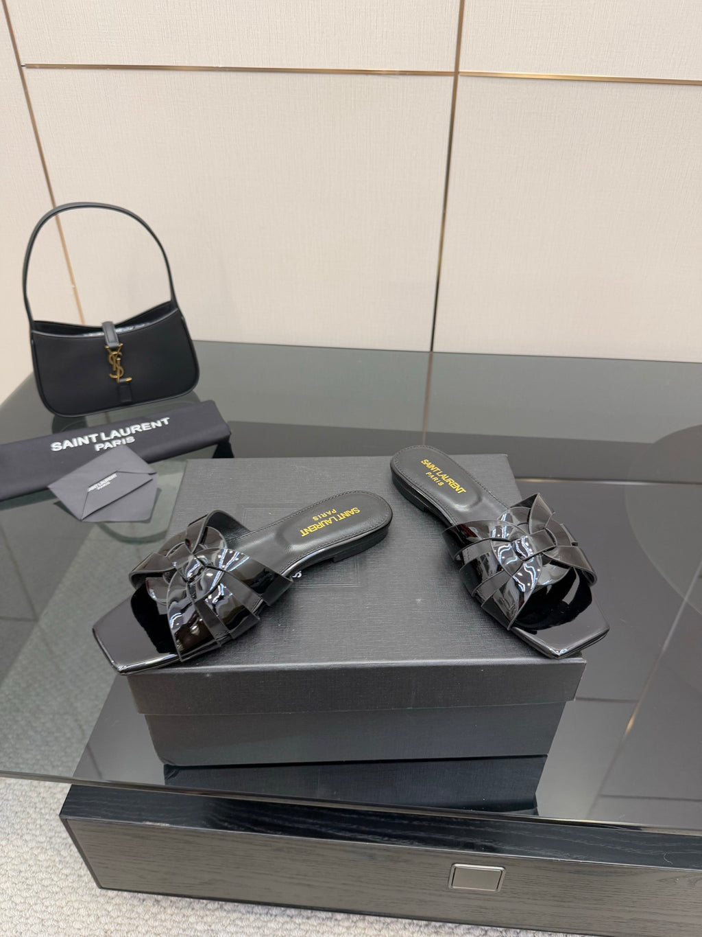 Black slide sandals with glossy straps and metallic buckle detail on a display box