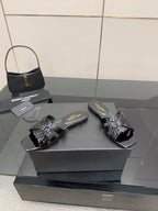 Black slide sandals with glossy straps and metallic buckle detail on a display box