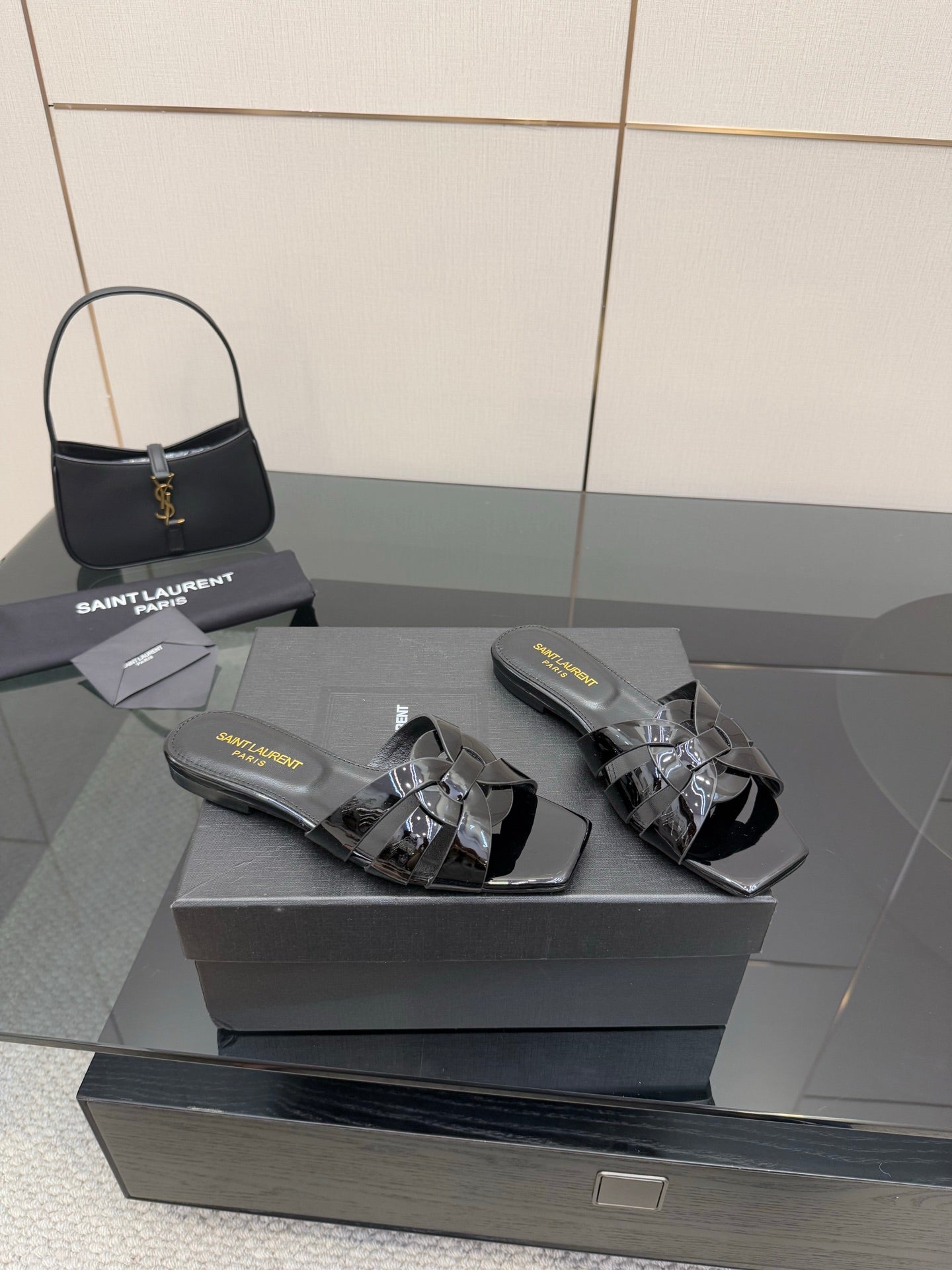 Black slide sandals on a box, patent straps with metallic buckle and shine detail