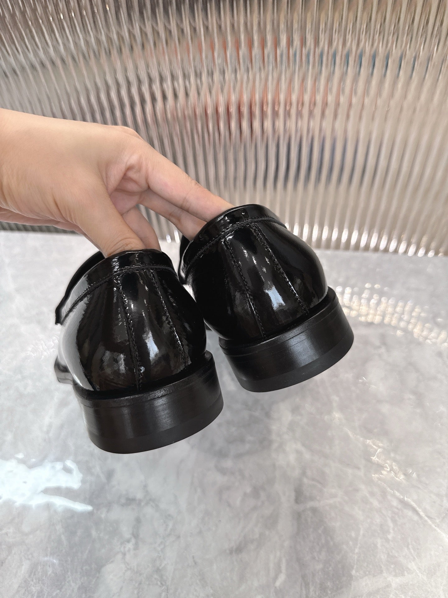 Hands holding black patent loafers, heel view showing low stacked sole.