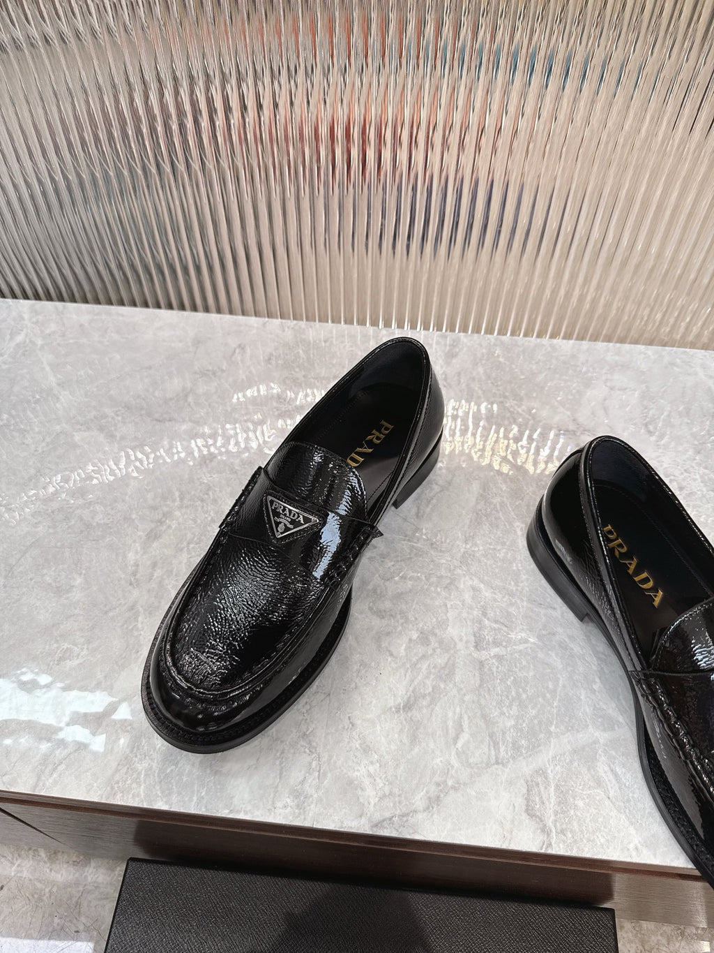Black patent loafers with Prada badge on the strap, angled front view.
