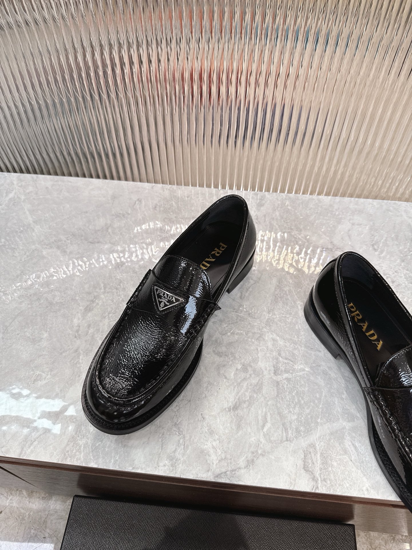 Black patent loafers with Prada badge on the strap, angled front view.