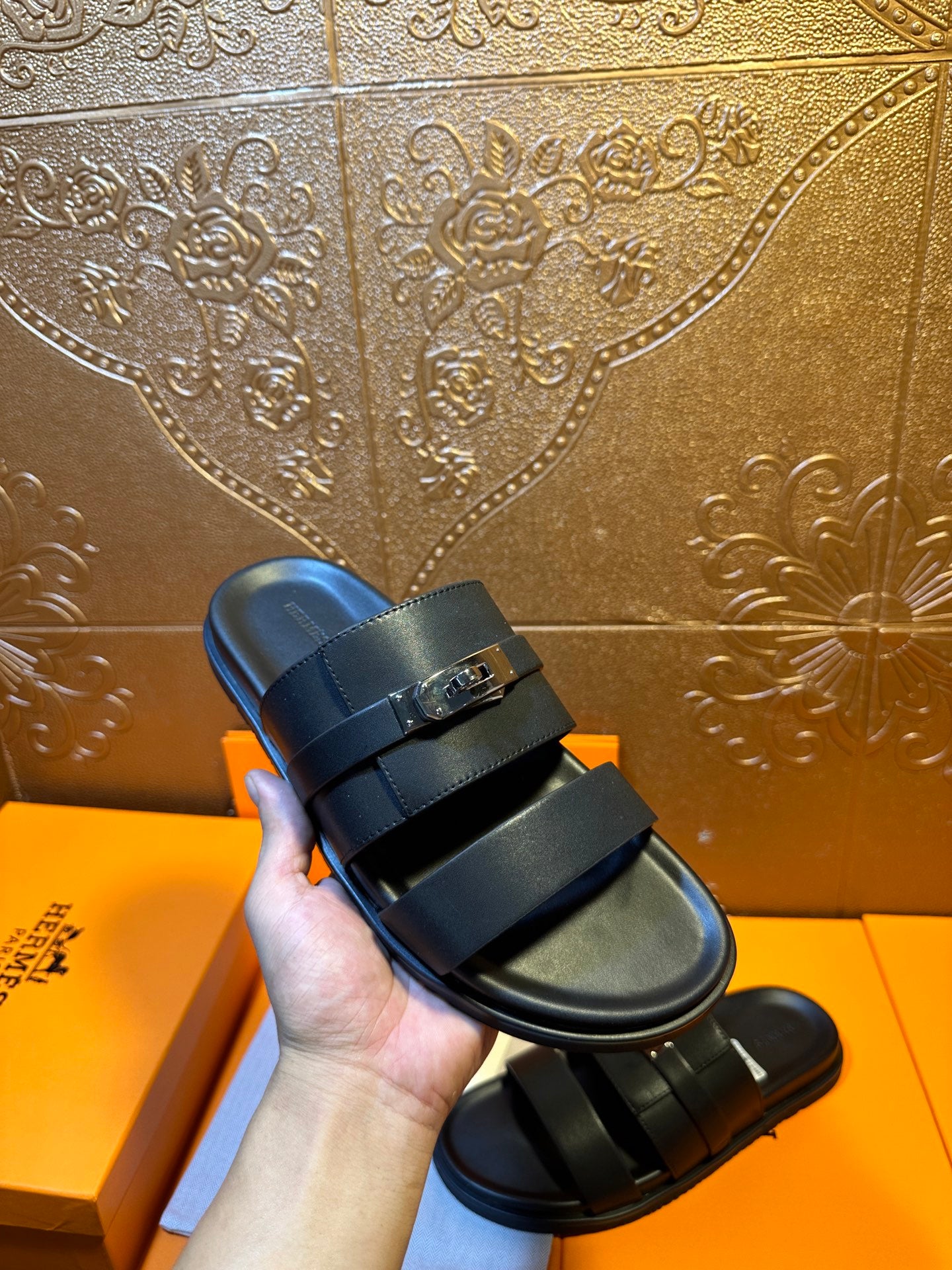 Hermes leather slide sandals held in hand, dark navy straps with gold-tone buckle