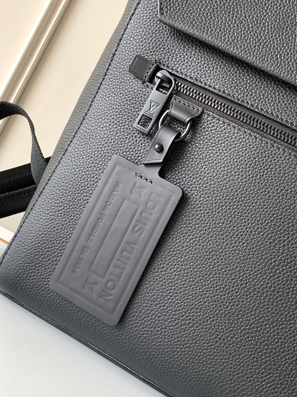 Men's Louis Vuitton bag close-up of front zipper pull and embossed leather patch, dark.
