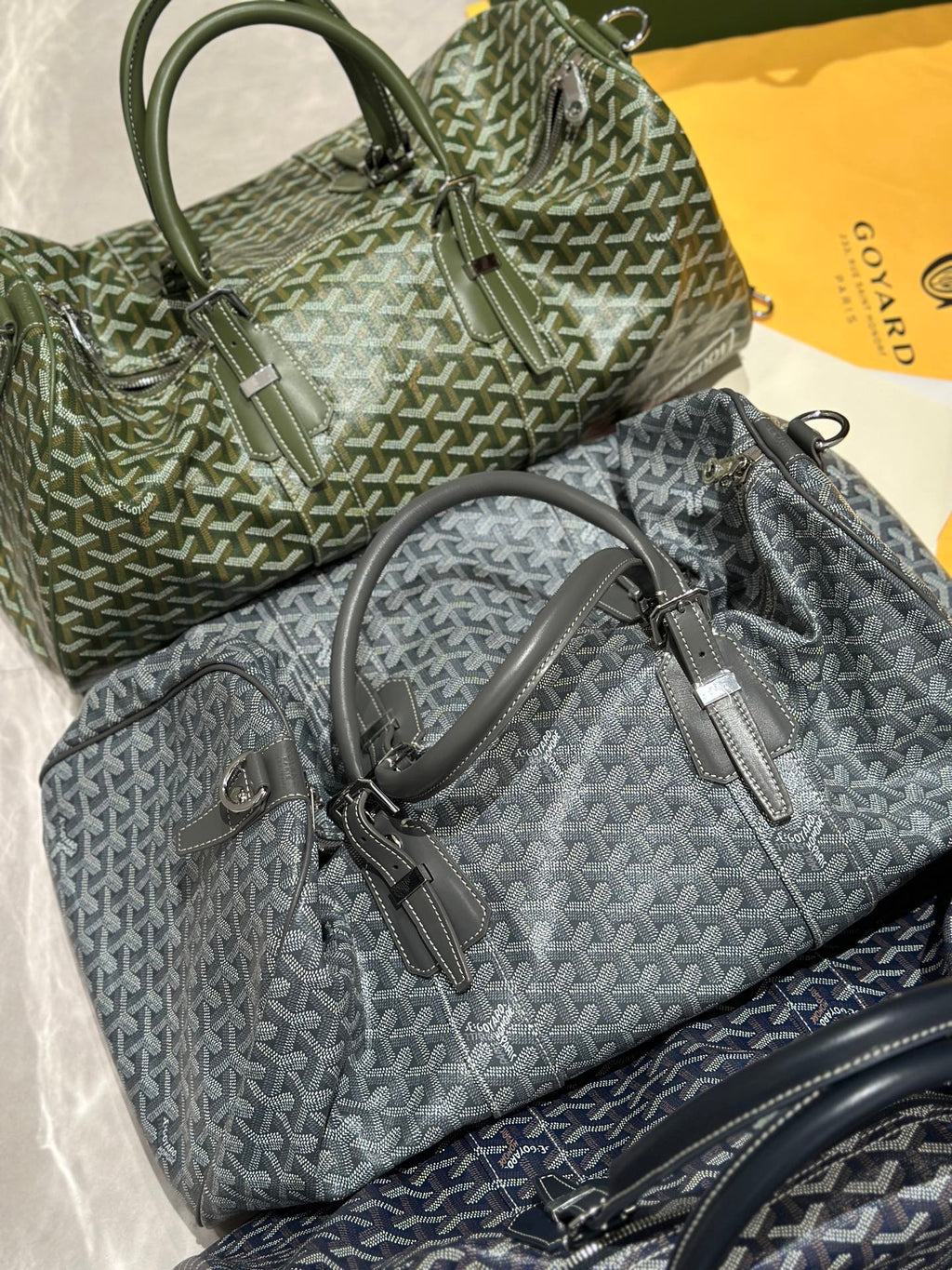 Stacked duffel bags in sage-green and dark-gray Goyardine with rolled top handles
