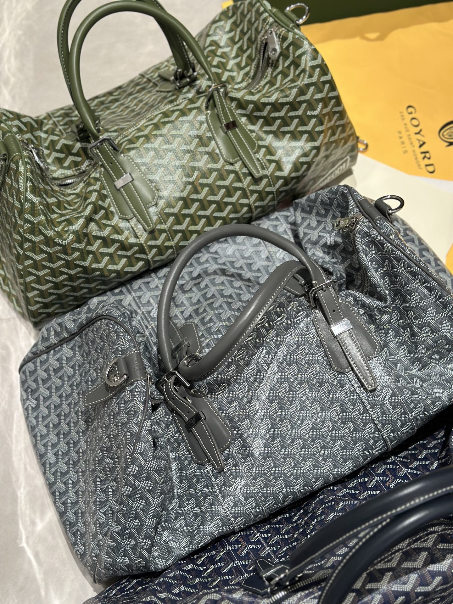 Stacked duffel bags in sage-green and dark-gray Goyardine with rolled top handles