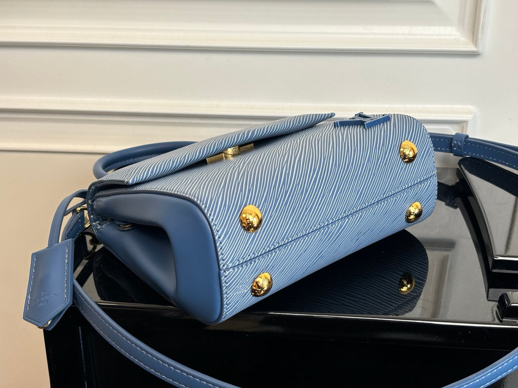 Blue textured handbag with flap, gold studs, and top handle on a flat base.
