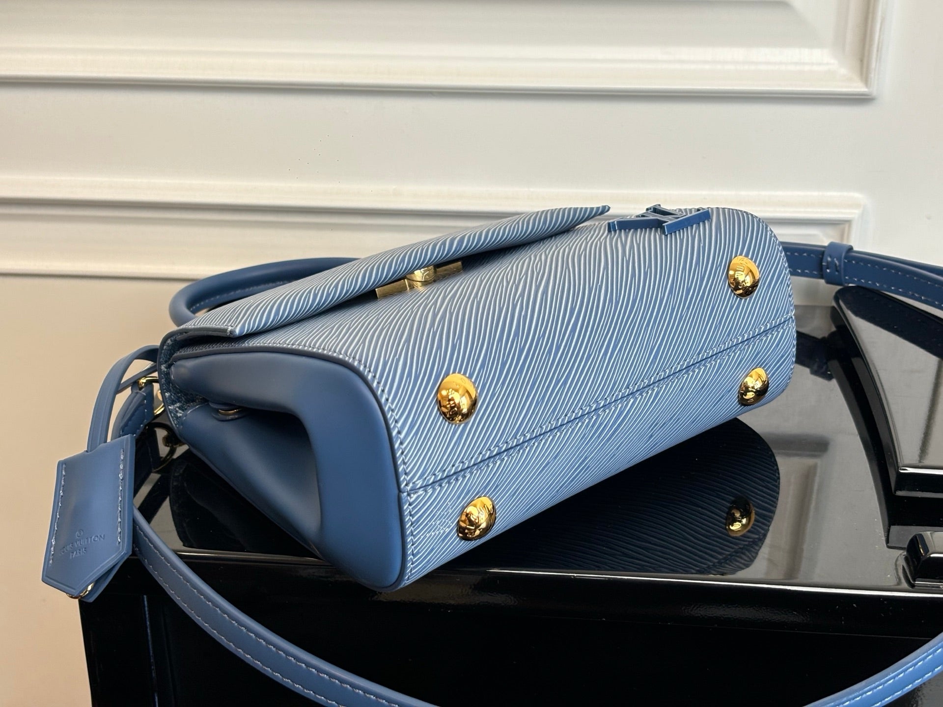 Blue textured handbag with flap, gold studs, and top handle on a flat base.