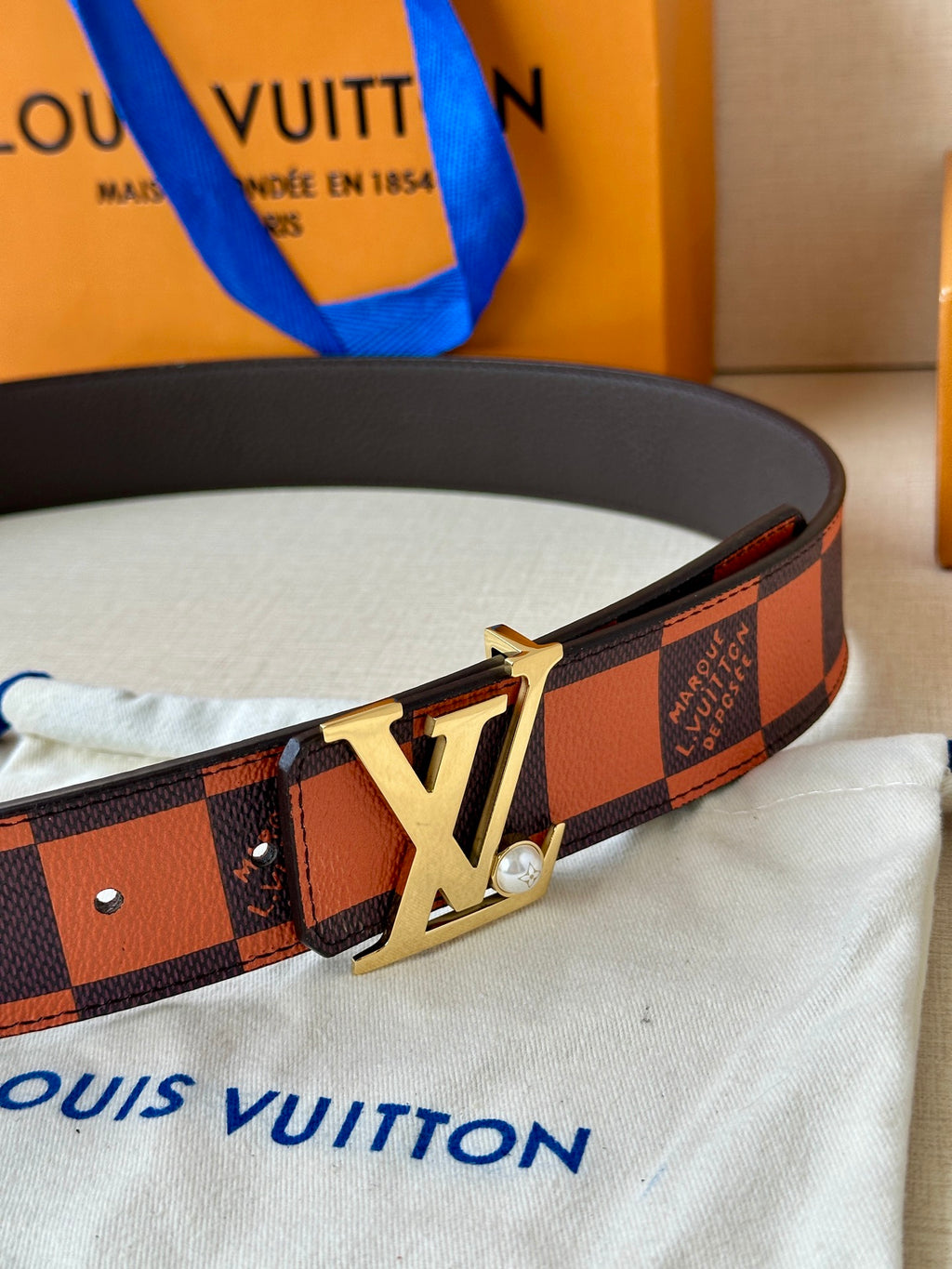 Louis Vuitton belt laid flat, orange-brown patterned strap and gold LV buckle.