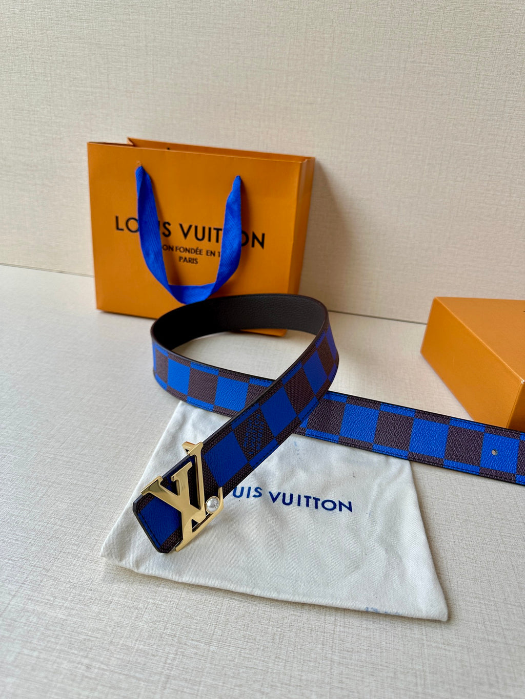 Louis Vuitton belt with blue check strap and gold-tone LV buckle on dark backing.