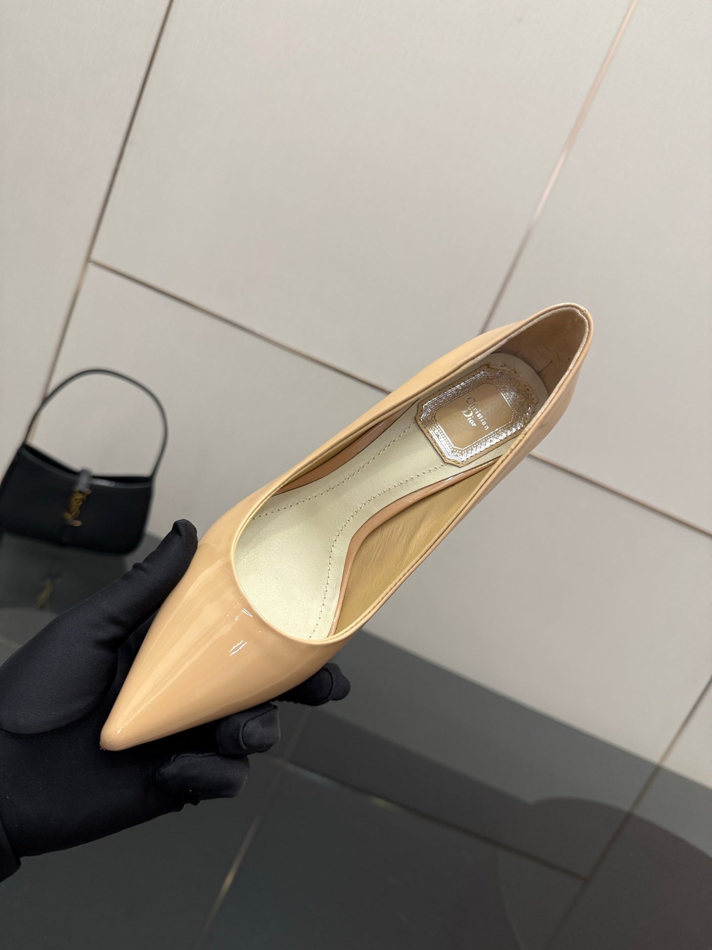 Beige patent pointed-toe pump with decorative strap, held in black glove