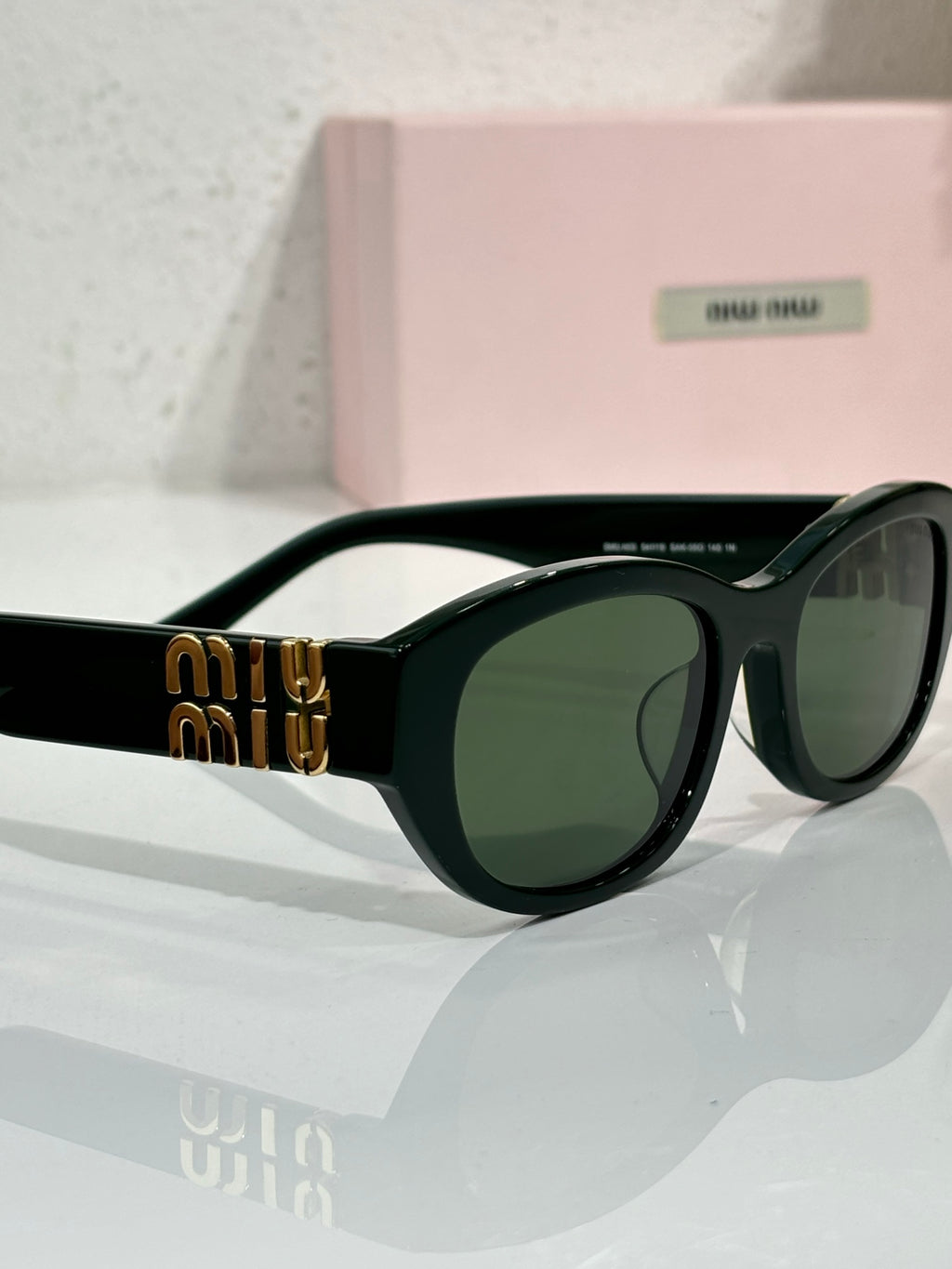 MiuMiu sunglasses close-up: black frame with green tinted lenses and gold temple logo