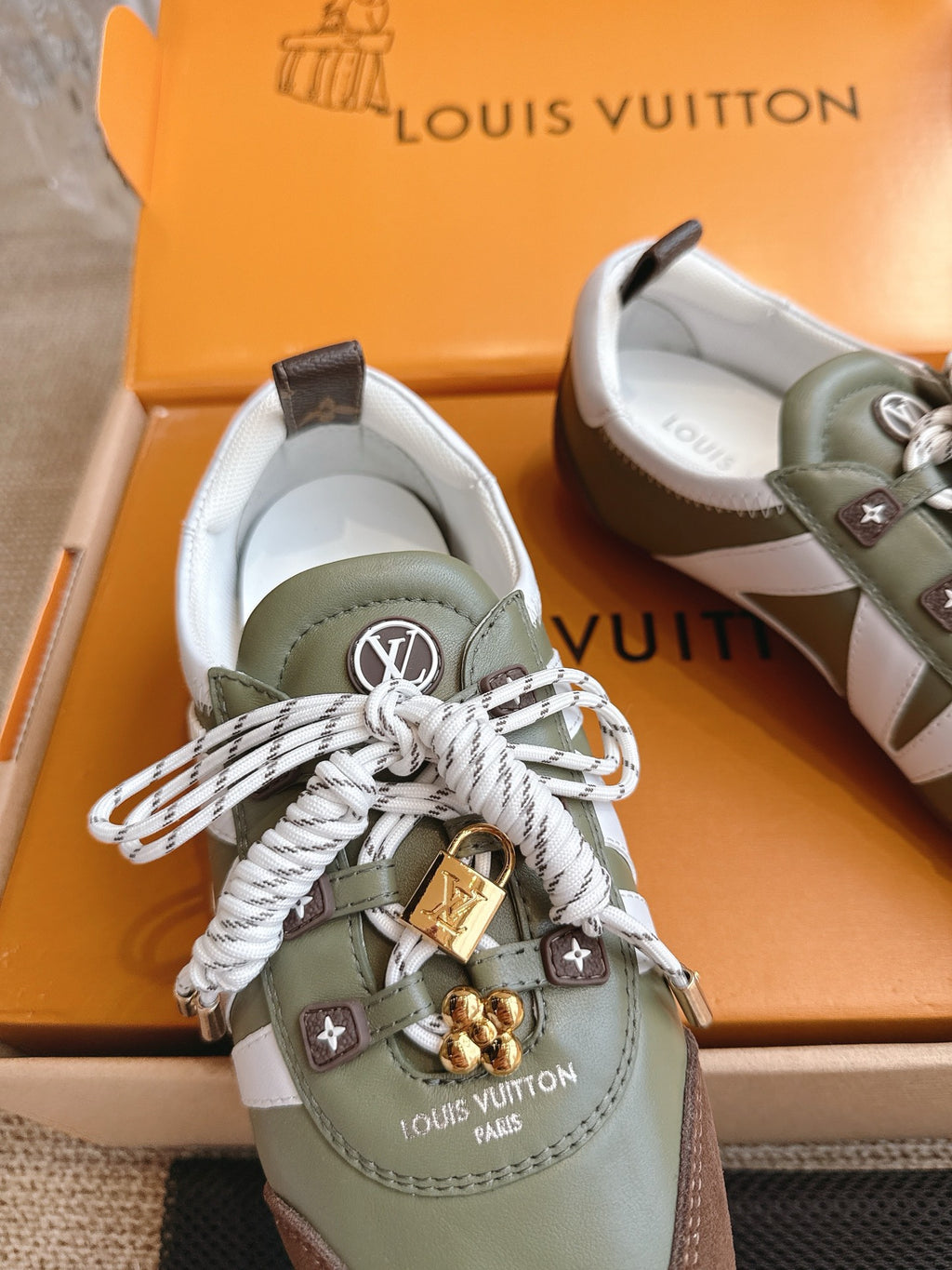 Close-up low-top sneaker, olive leather with brown heel, white laces and gold charm.
