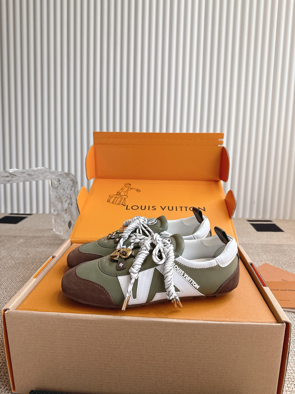 Low-top sneakers in open box, olive and brown uppers with white laces and charms.