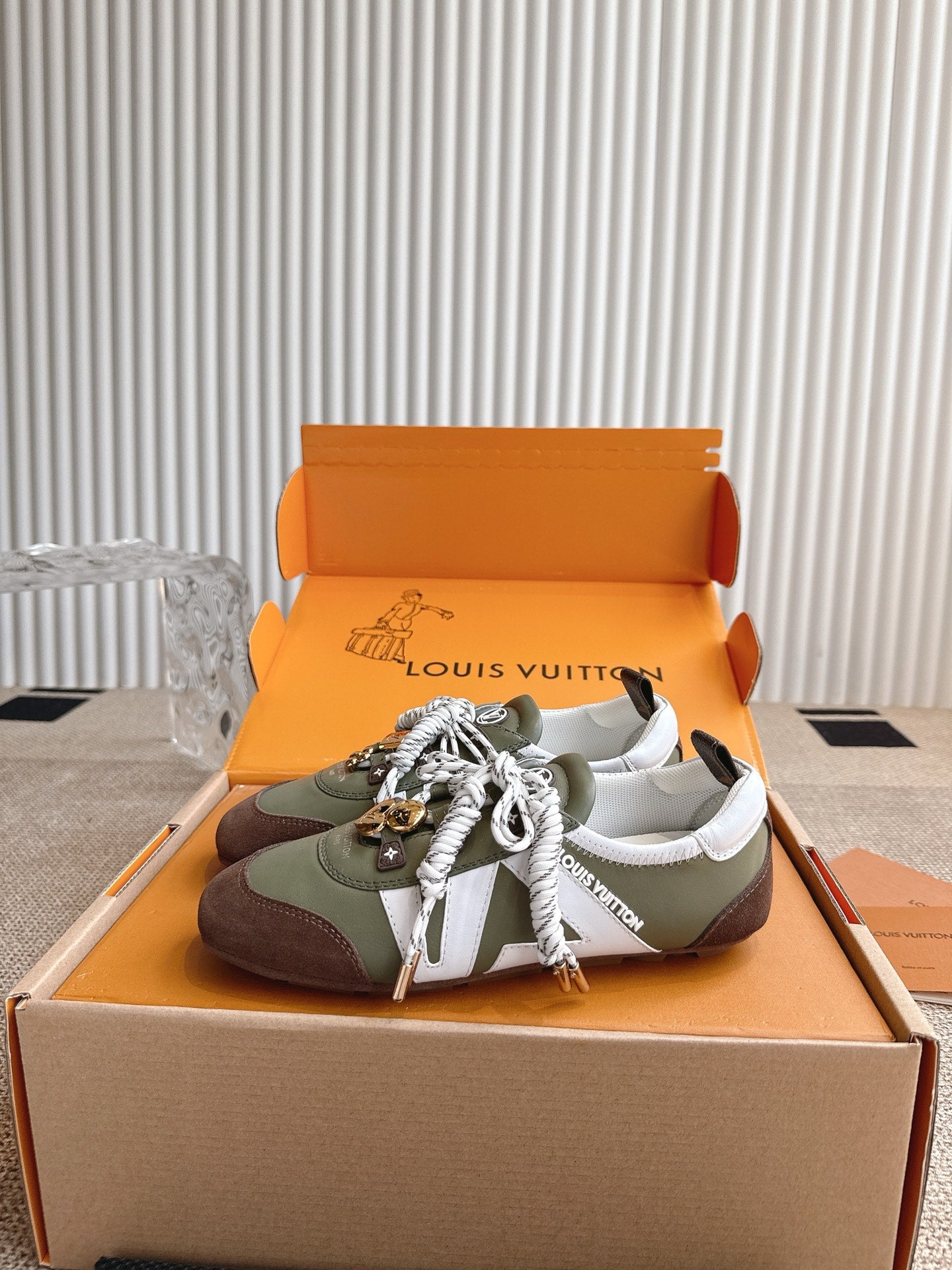 Low-top sneakers in open box, olive and brown uppers with white laces and charms.