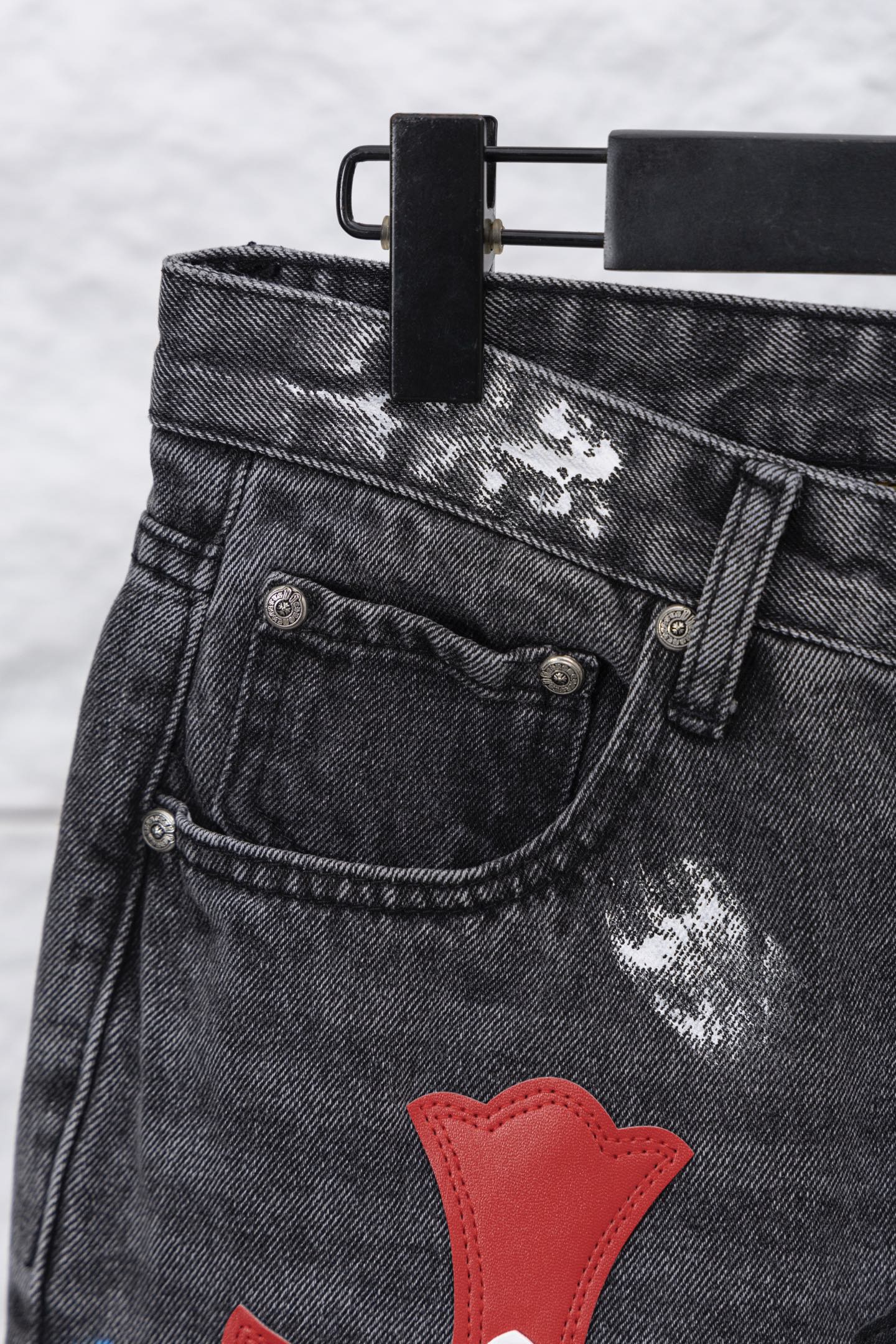 Black denim pants waistband and front pocket with red heart-like cross applique; close-up.