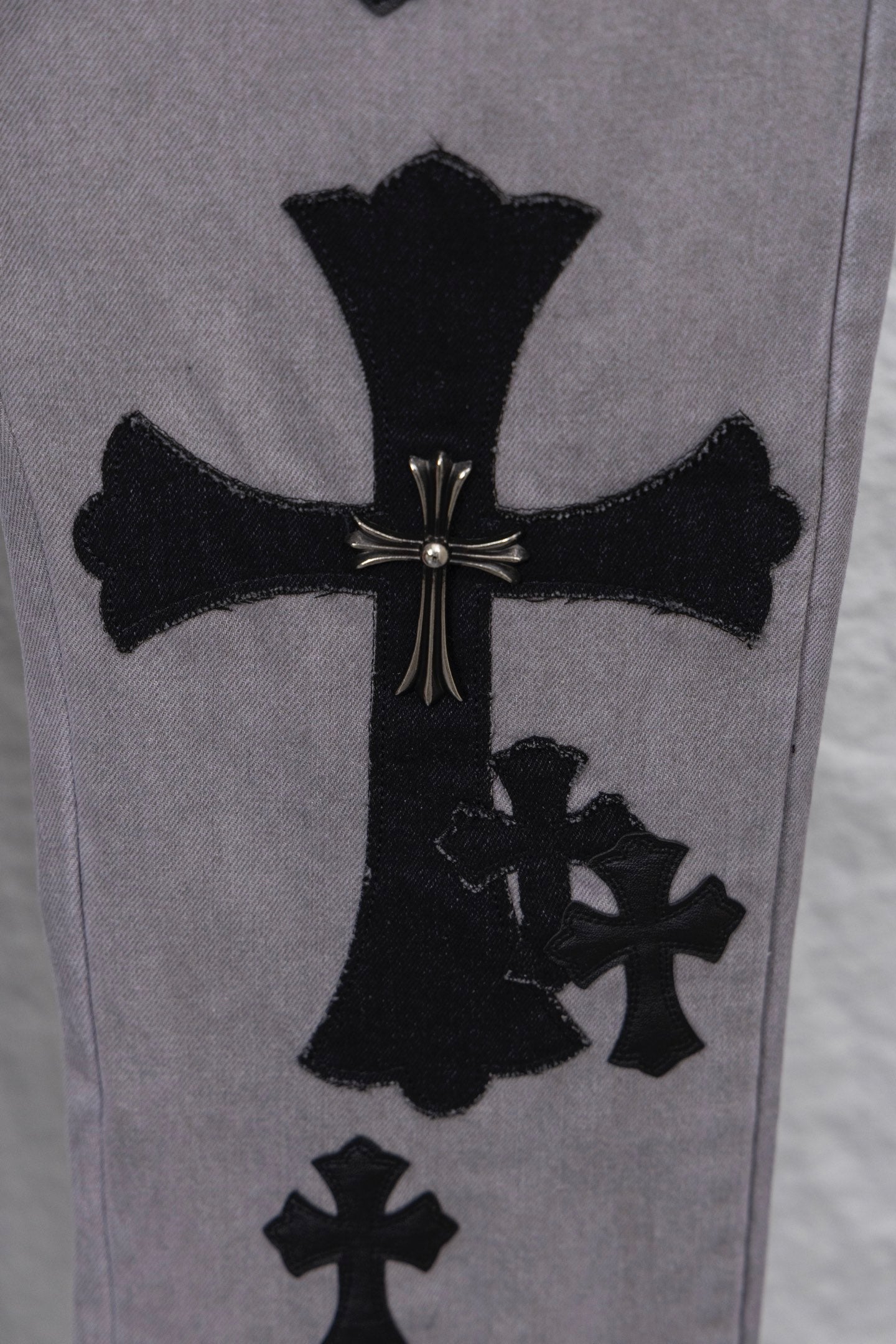 Light gray denim pants with large black cross applique and smaller cross motifs, close-up