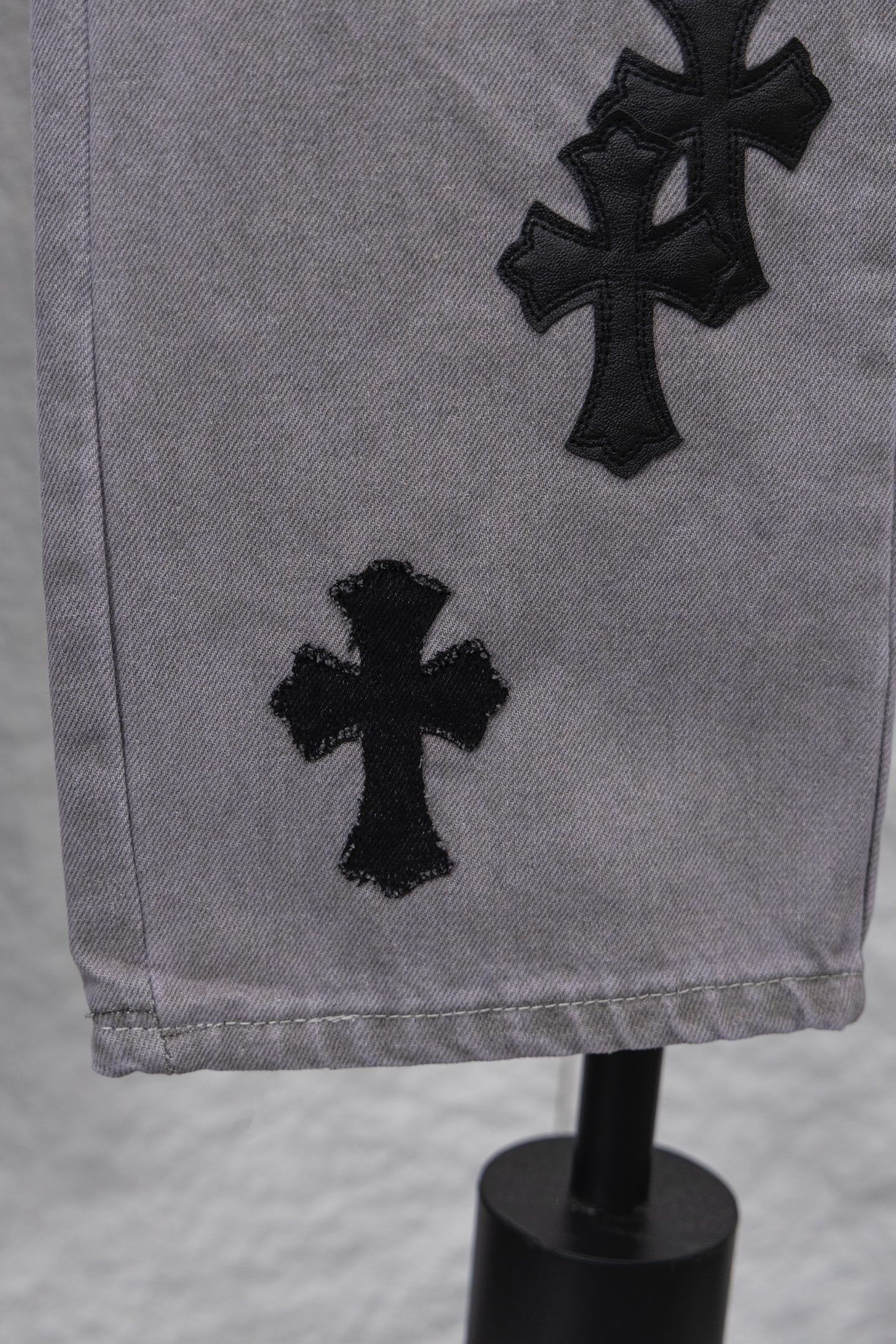 Light gray denim pants with black cross applique near hem, close-up