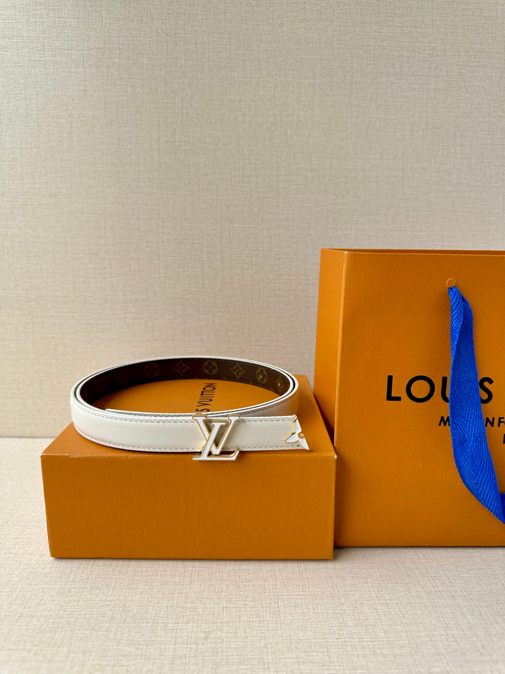 White leather belt with gold-tone LV buckle over dark monogram strap.
