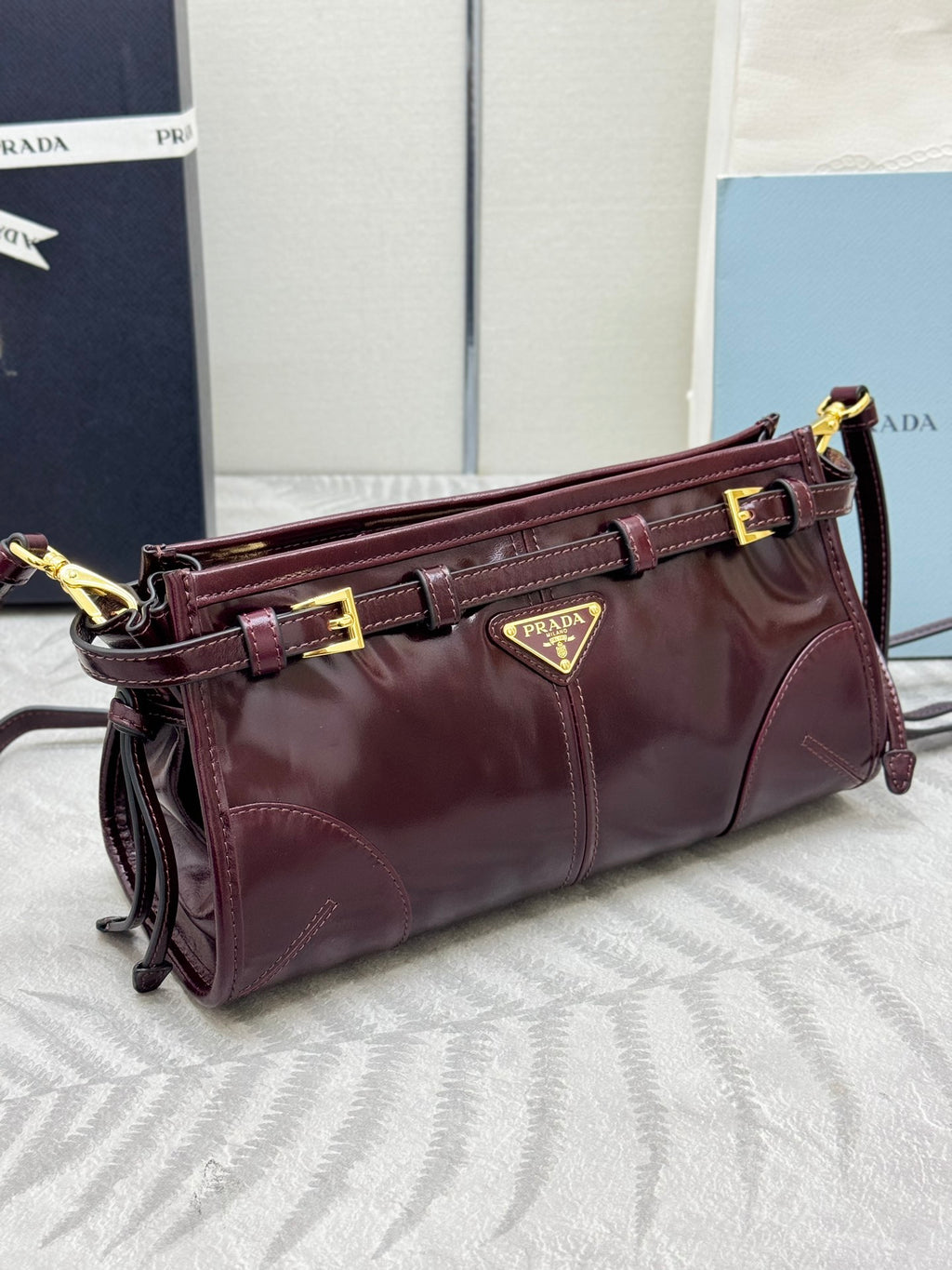 Prada burgundy leather shoulder bag with side straps, gold buckle details, and logo plaque
