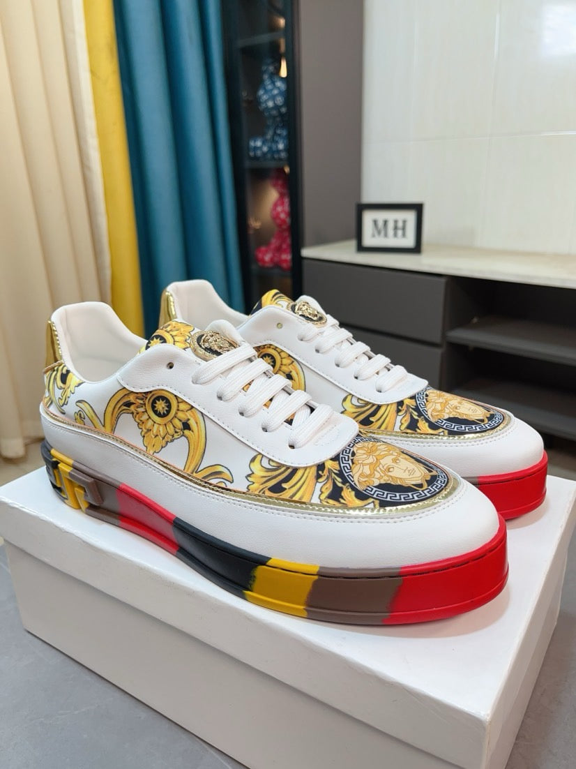 White luxury sneakers with gold baroque print, white laces, and red/yellow platform sole.
