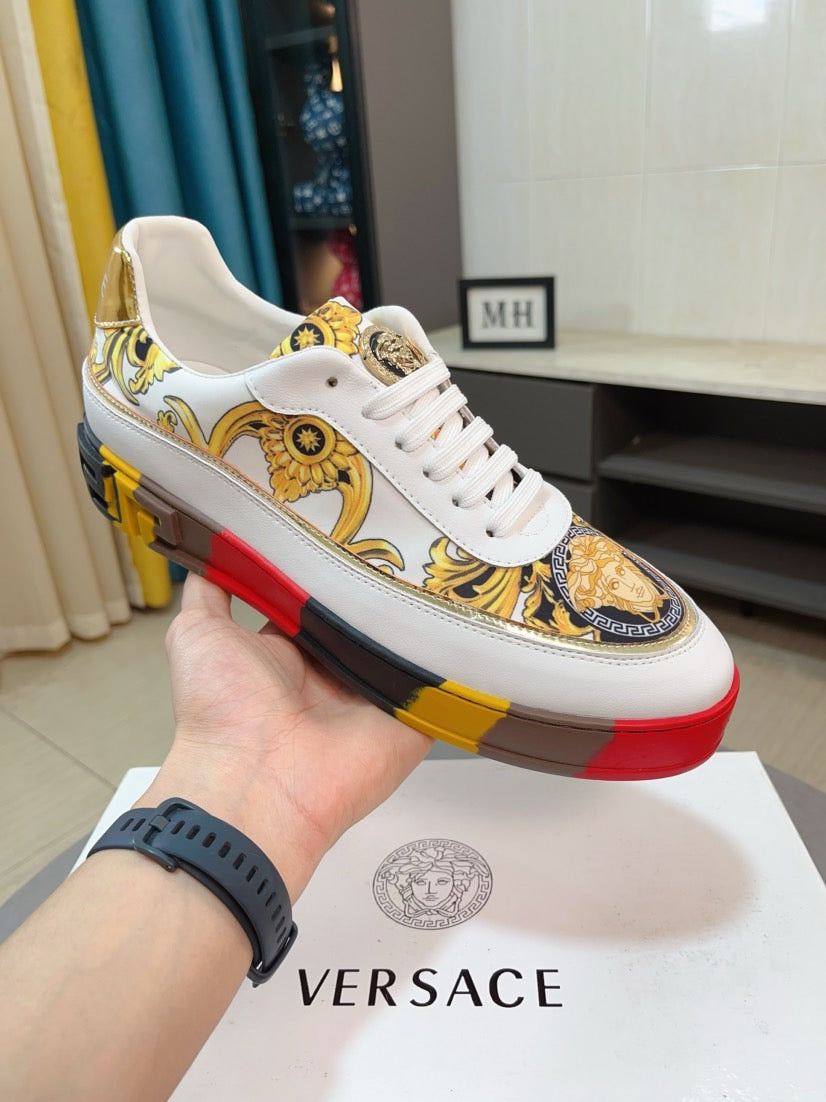 White luxury sneakers with gold baroque pattern, gold lace tab, and red/yellow/black sole.