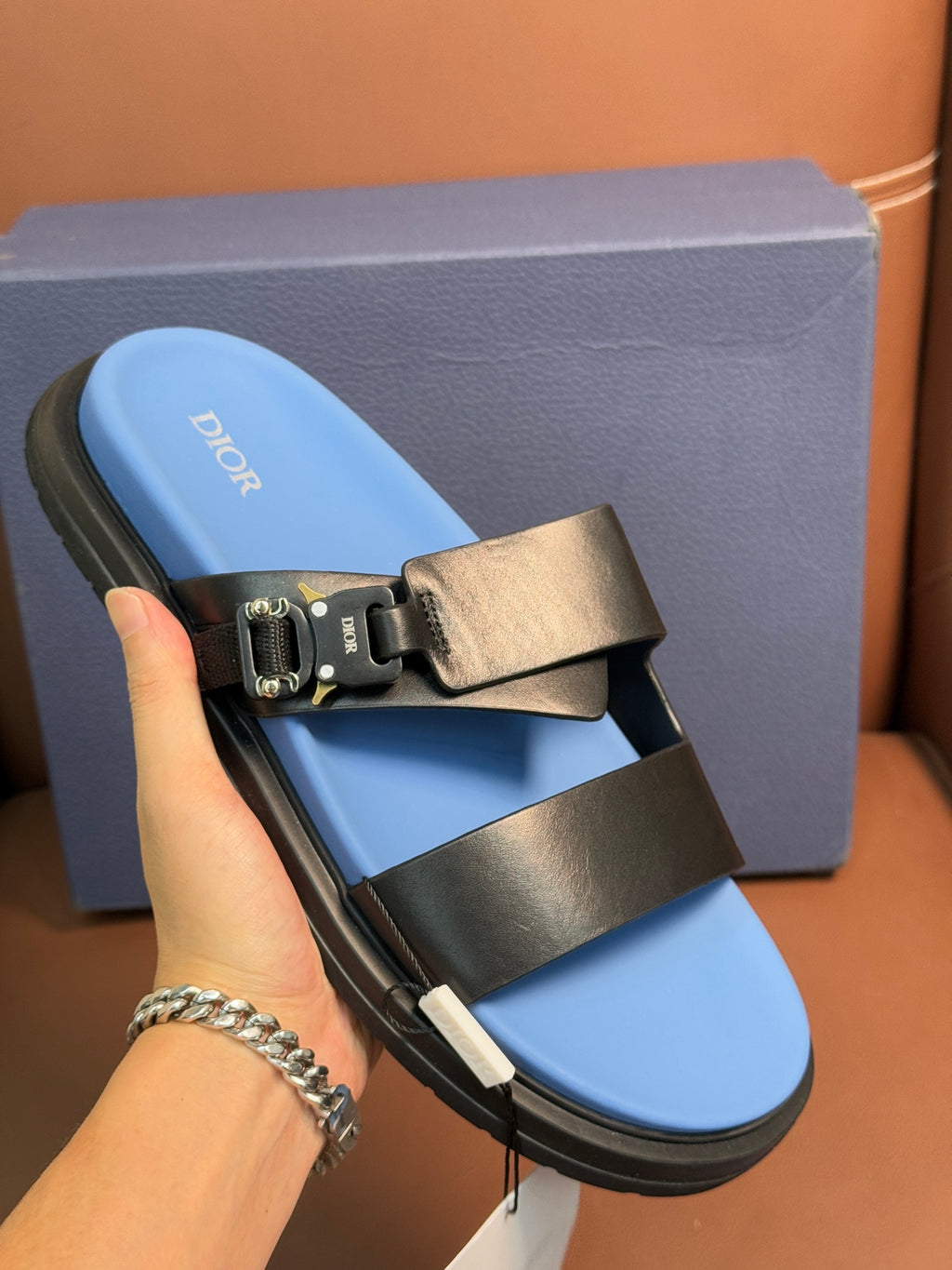 Dior slide sandals shown in hand, blue footbed with black straps and gold buckle