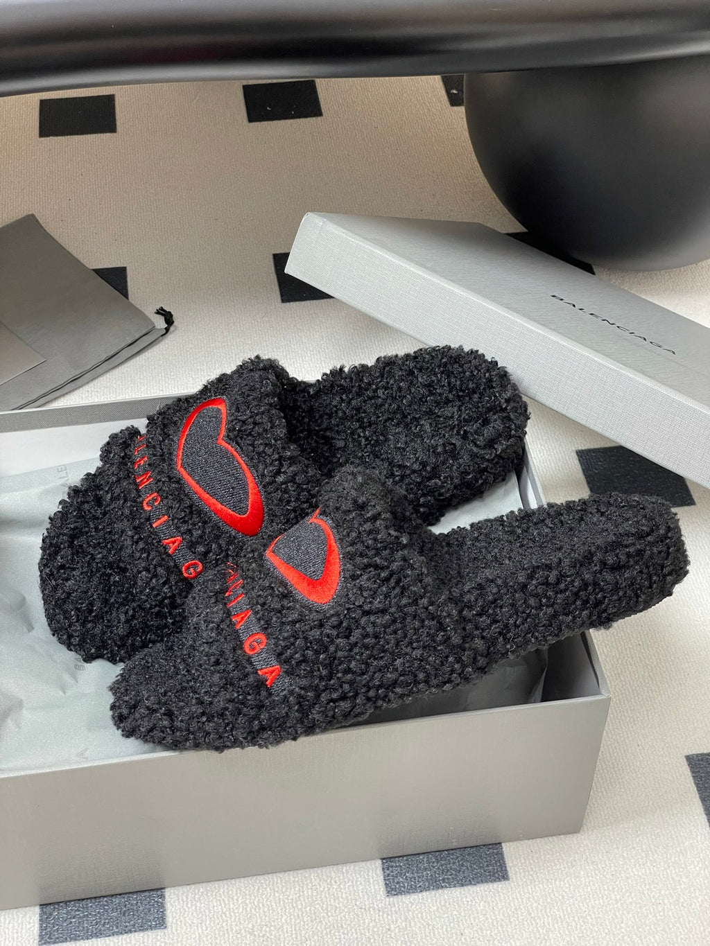 Black teddy-fleece slide sandals with red heart logos in an open shoebox