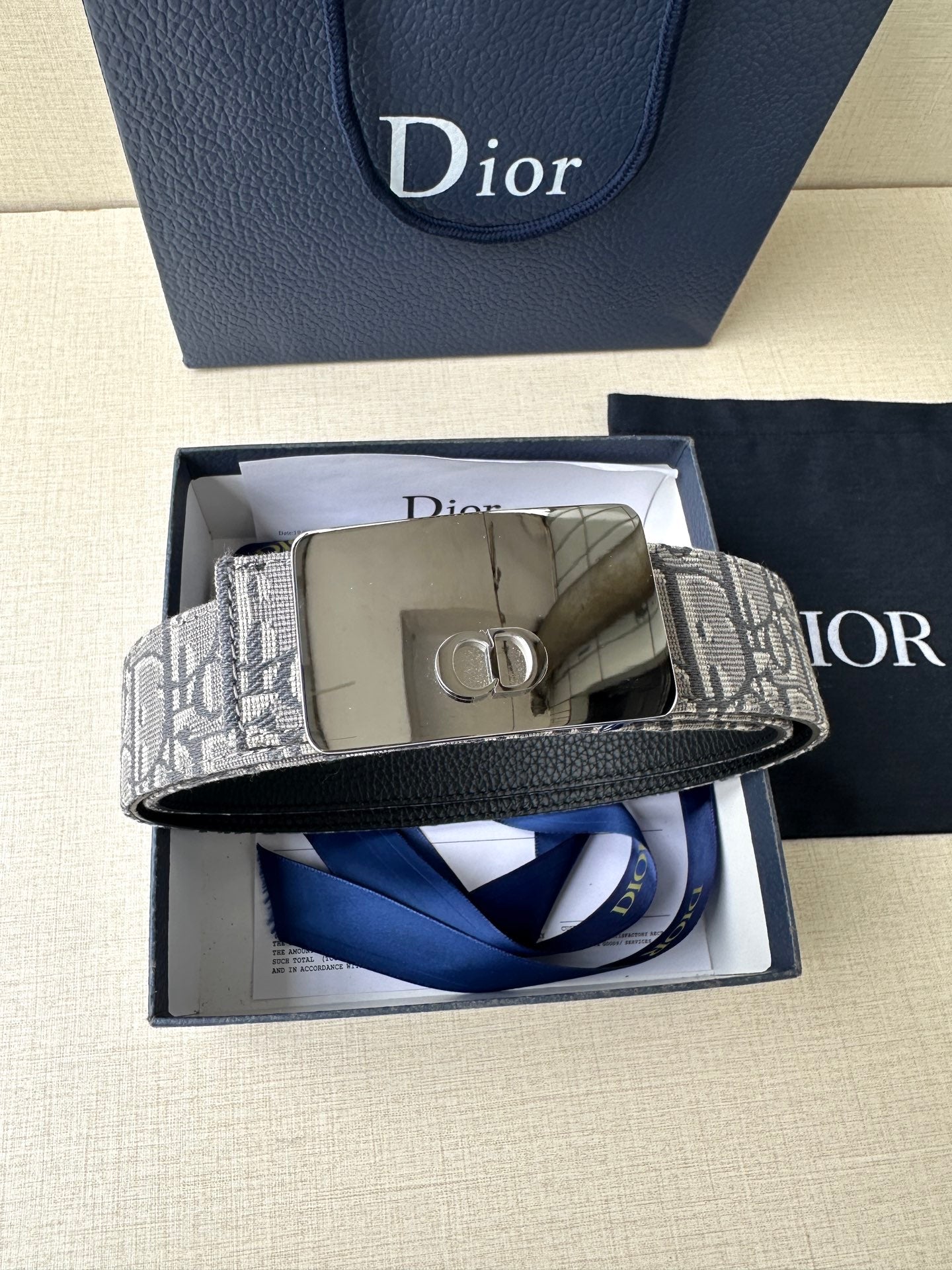 Dior belt with gray monogram strap and gold buckle, laid across box inserts