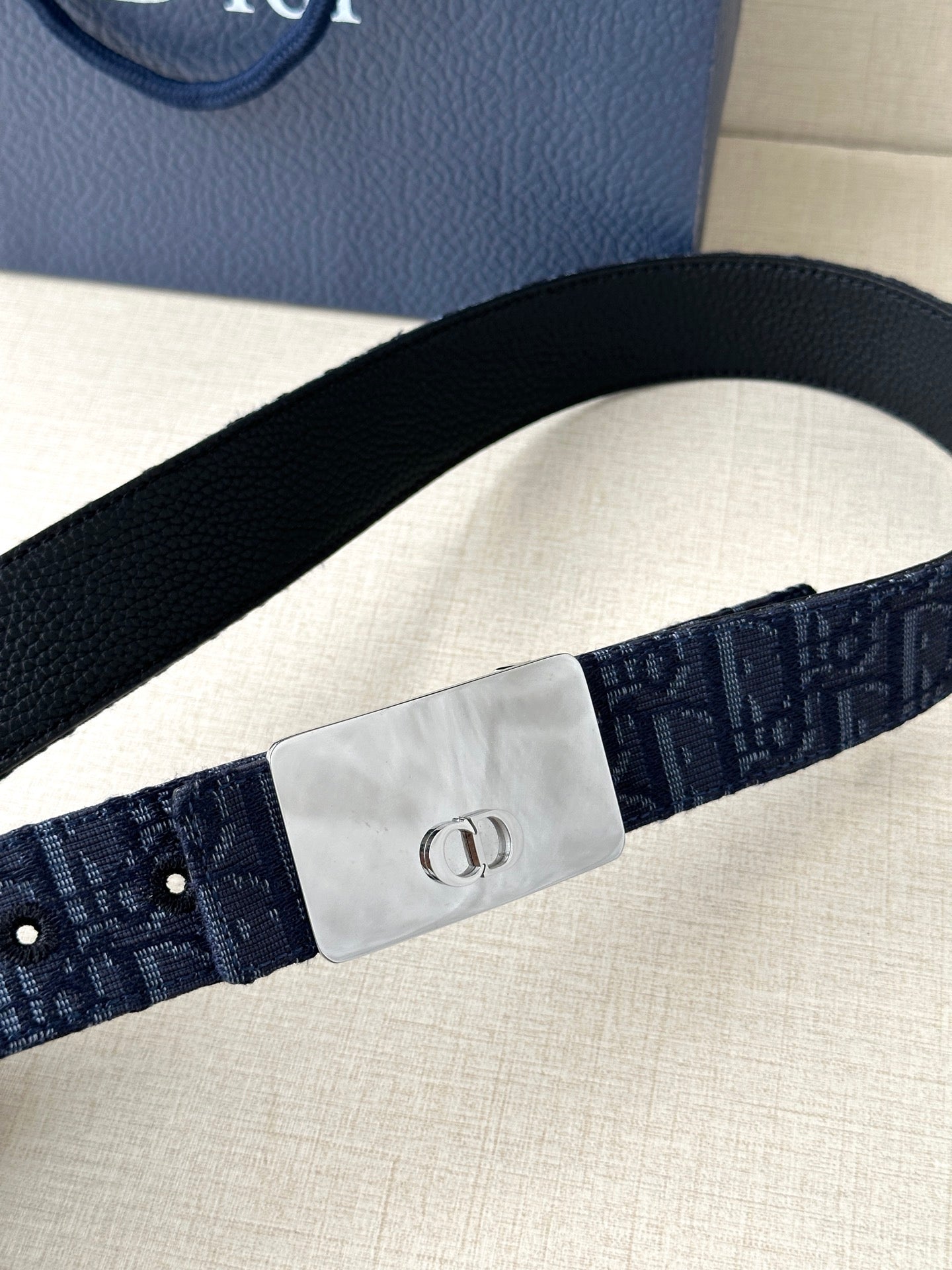 Dior belt close-up: navy patterned strap with silver buckle at center and punched holes.