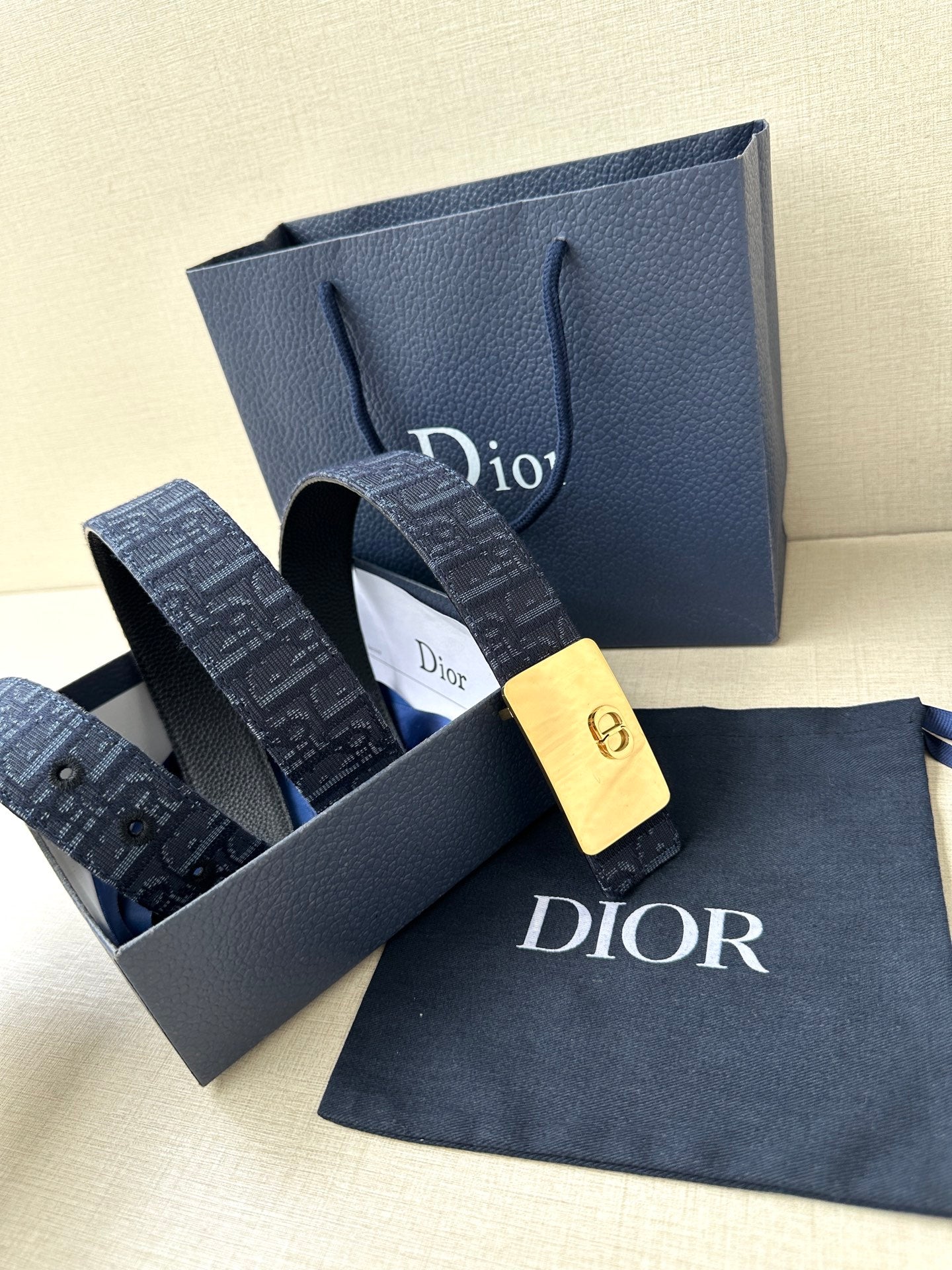 Dior belt with navy patterned strap and gold-tone buckle plaque, beside gift box and bag.