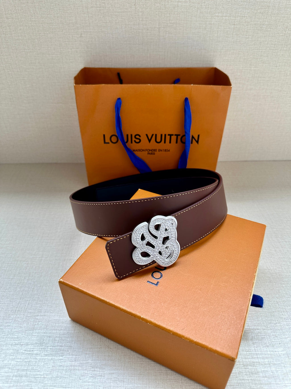 Dark brown leather belt with silver-tone logo buckle, shown on display box