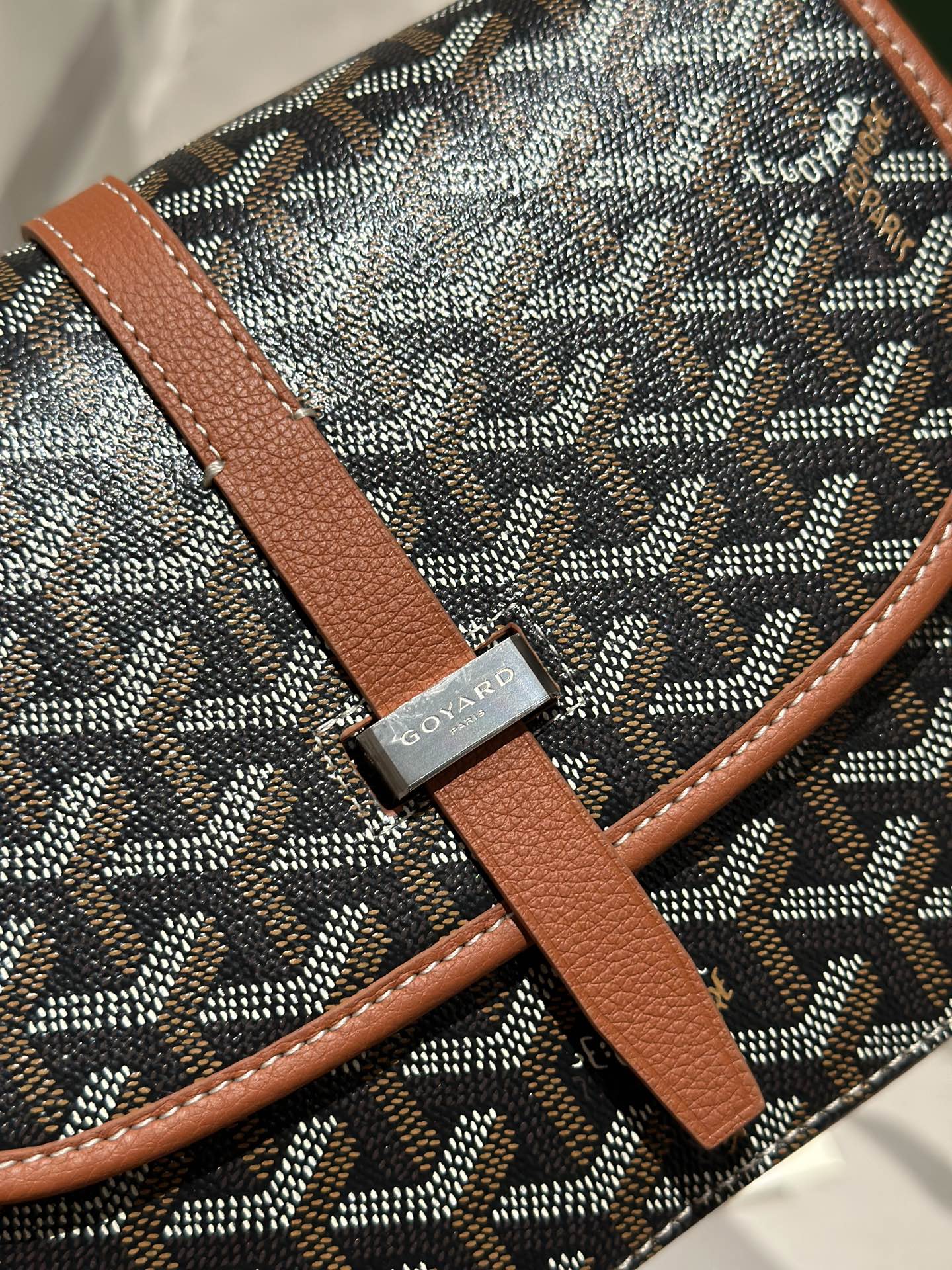 Goyard logo leather patch on tan strap over gray patterned canvas bag.