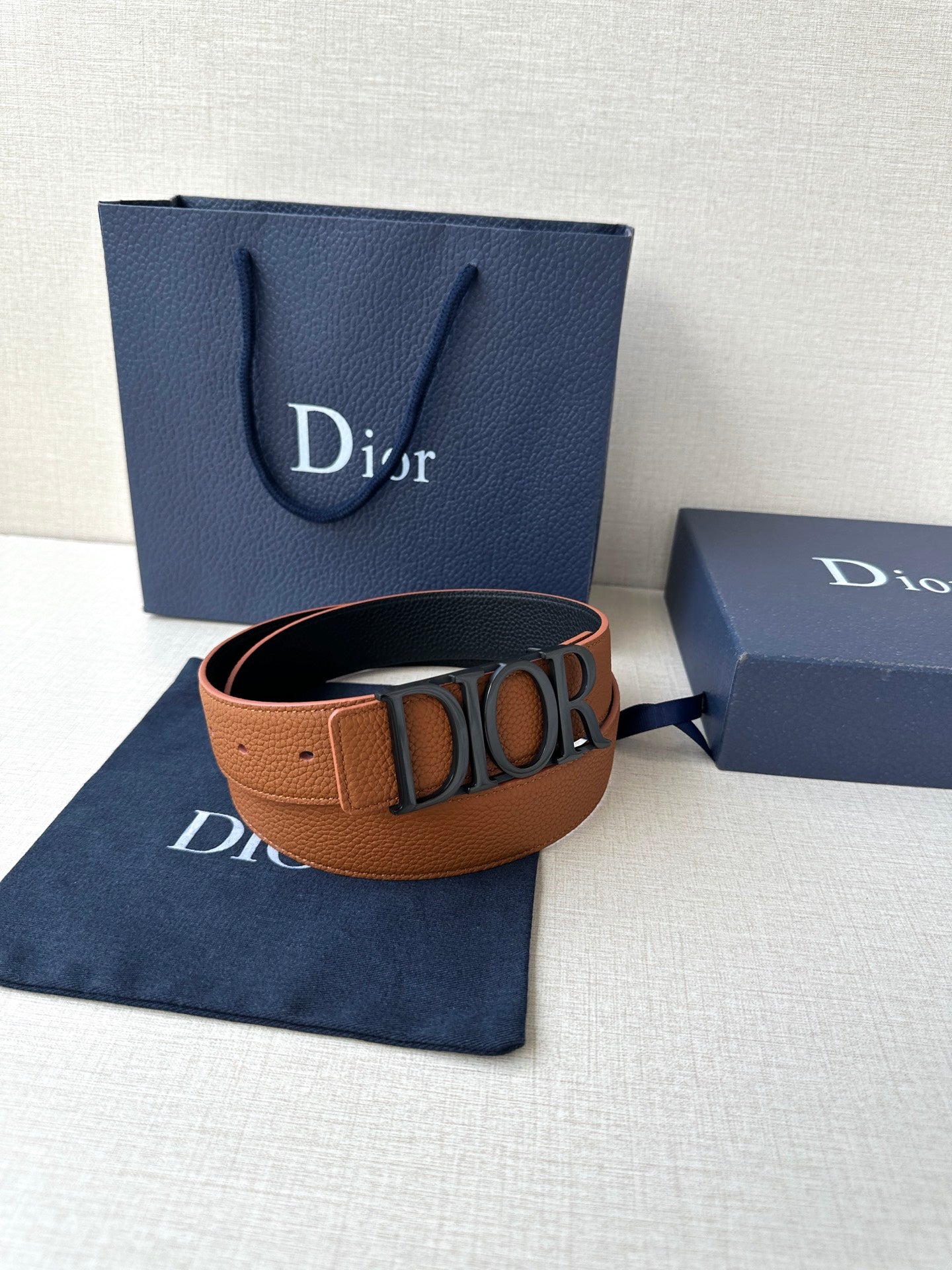 Tan leather Dior belt with engraved buckle, resting on folded blue dust bag.