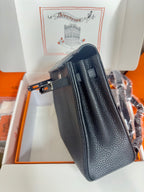 Hermes handbag side view in dark textured leather with top handle and belt strap.