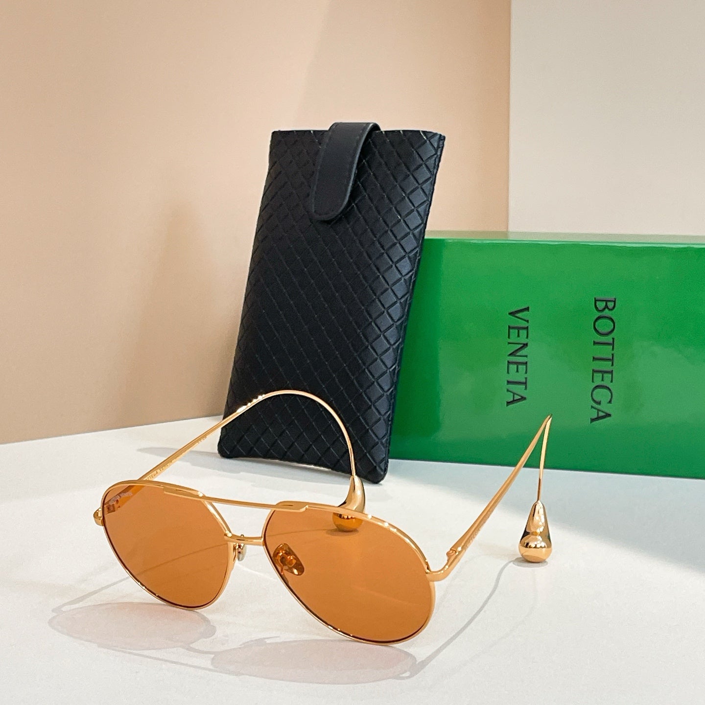 Bottega Veneta glasses with gold frame and amber-tinted lenses, beside black case