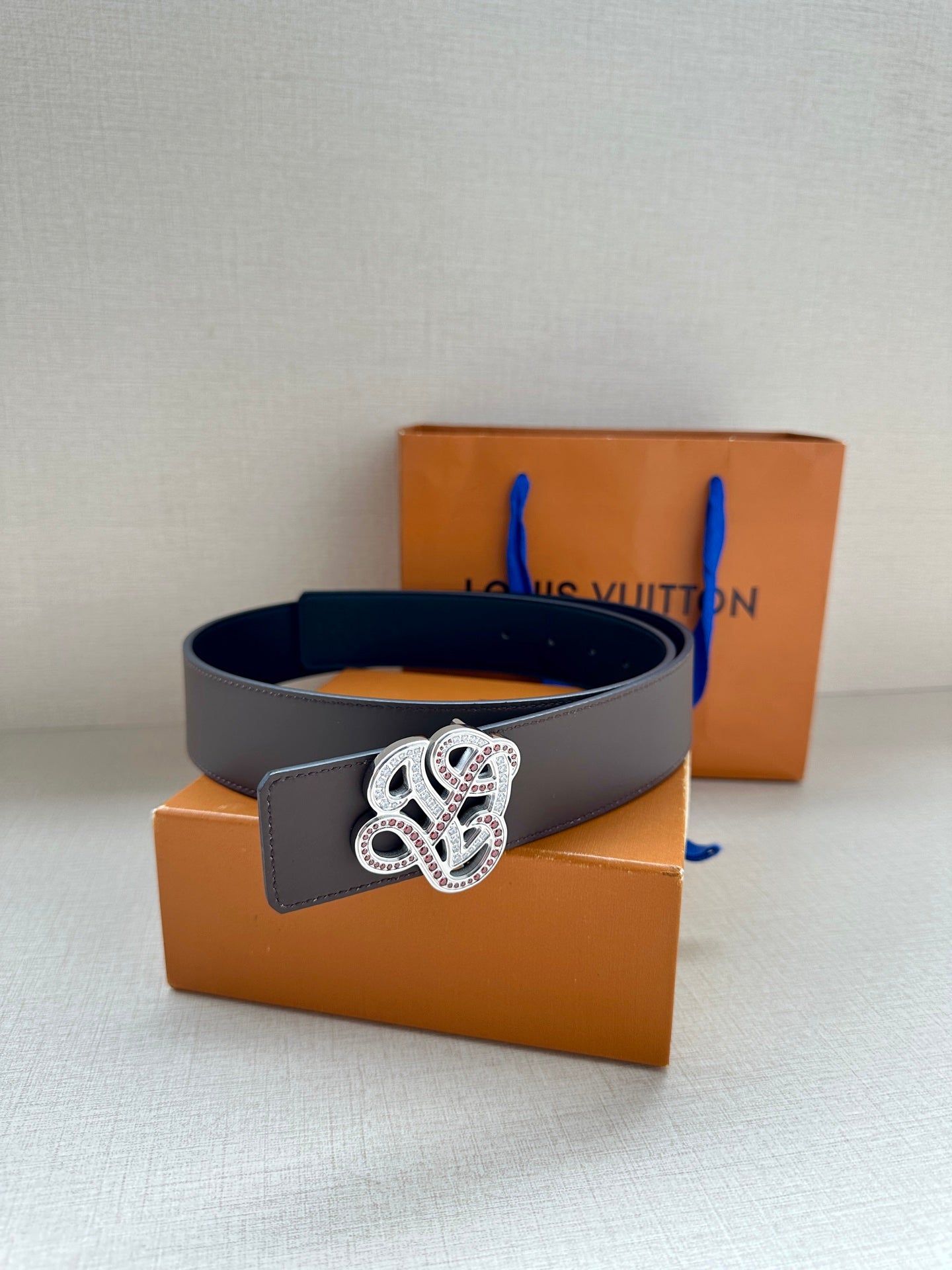 Dark navy leather belt with white-and-pink gem monogram buckle over stacked boxes