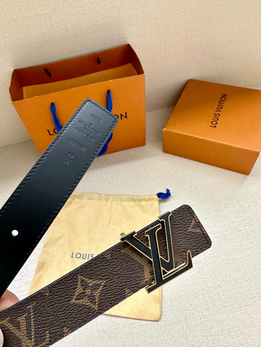 Monogram belt with gold-tone LV buckle, dark canvas strap and blue tag detail.