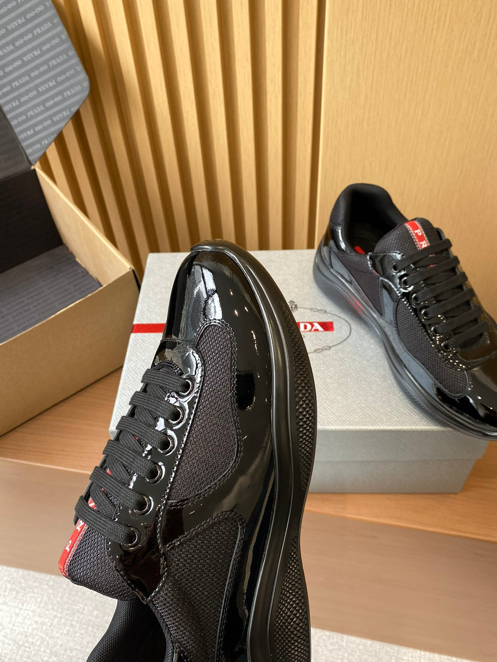 Black Prada lace-up sneakers with perforated side panels and red logo tab, on box.
